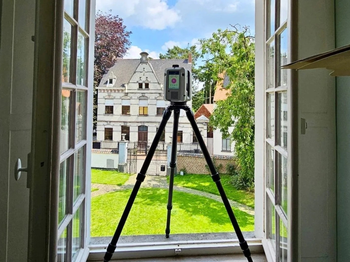 Photo of a building seen through an open window, with a black tripod and a device mounted on top, in a room with white walls and a small shelf to the right.