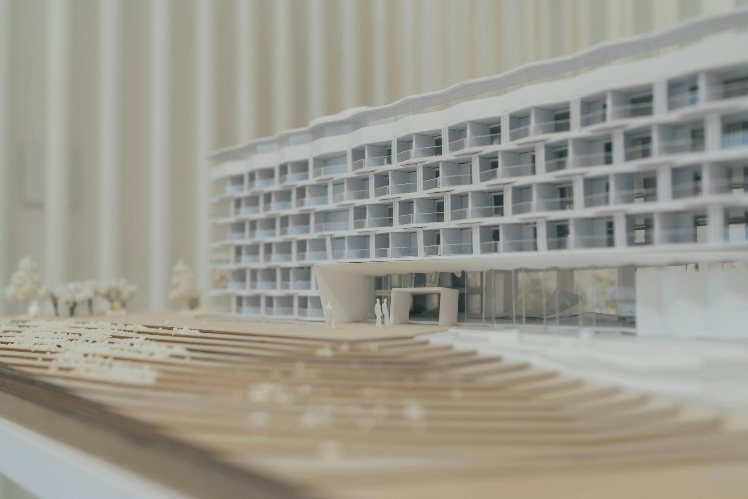 Architectural model of a modern, multi-story building with curved facade and glass balconies, featuring miniature figures near the entrance.