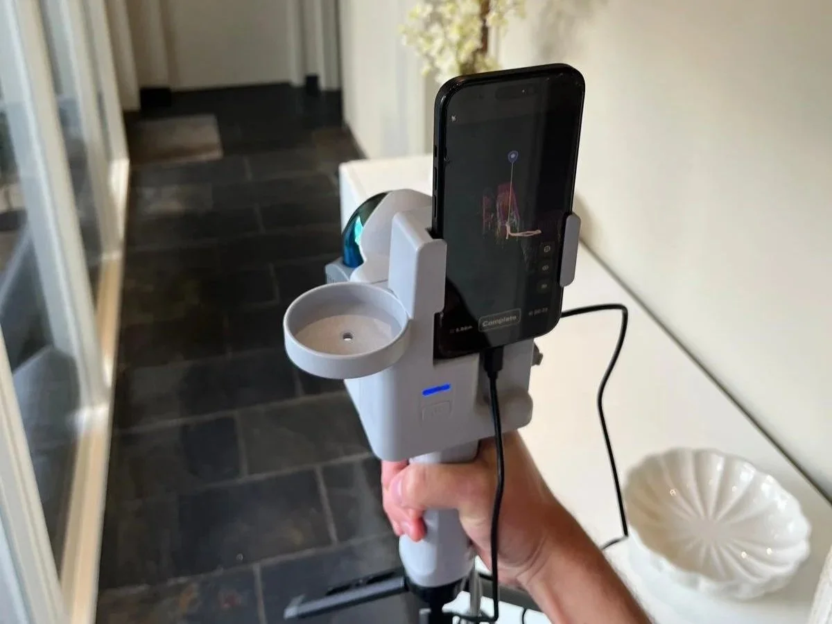 A person holding a handheld device with a smartphone mounted on it, capturing an image of a chair with a colorful abstract design, in a room with tiled floor and shelves on the left.