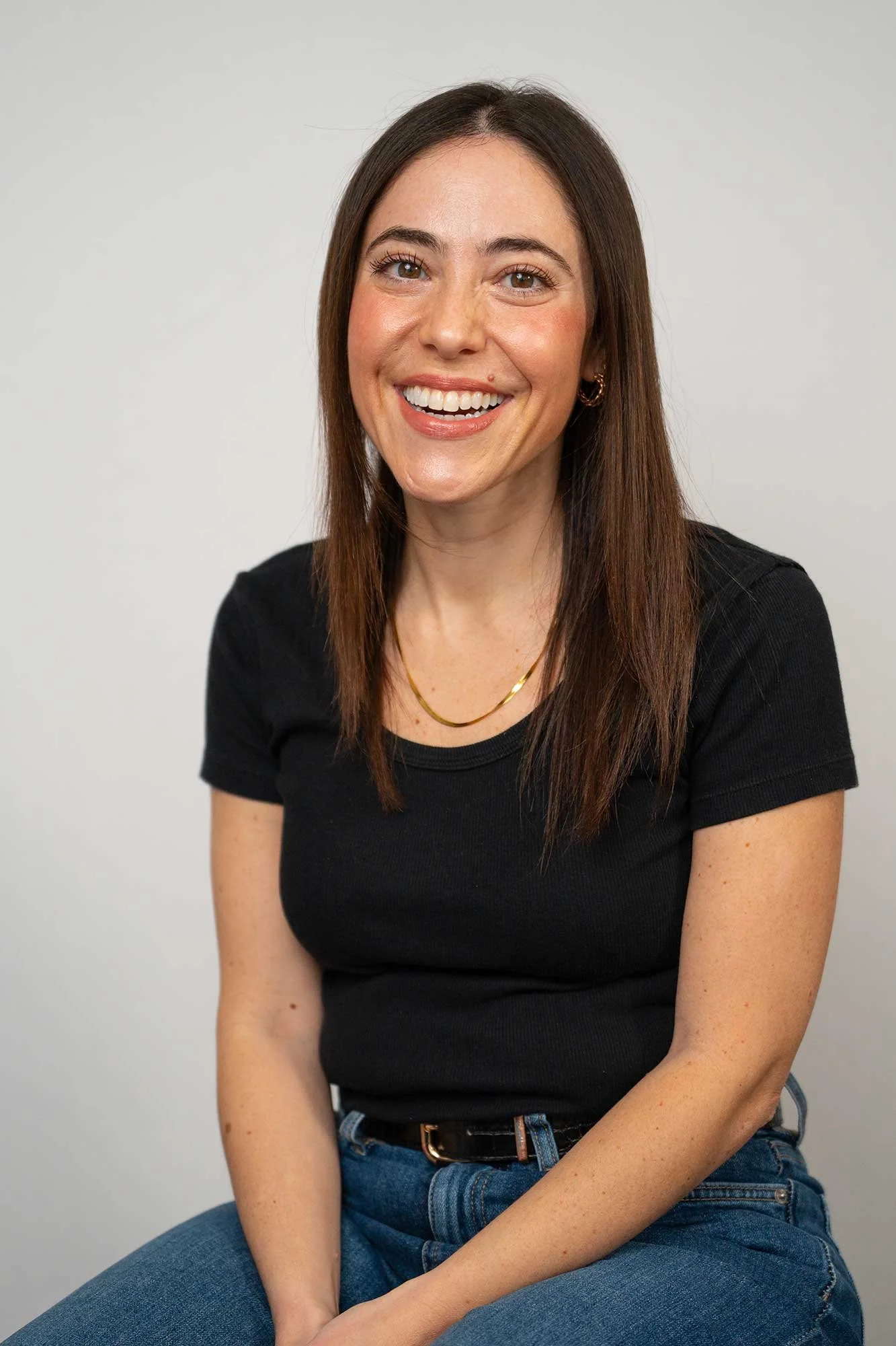An experienced London therapist with long dark hair wearing a black t-shirt and blue jeans, smiling and sitting against a plain light gray background.