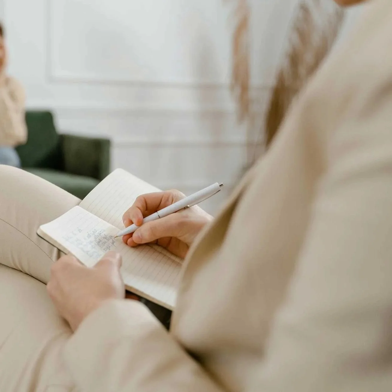 Close-up of a therapist in a beige outfit sitting on a cream-colored sofa, holding a small notebook and writing with a white pen.