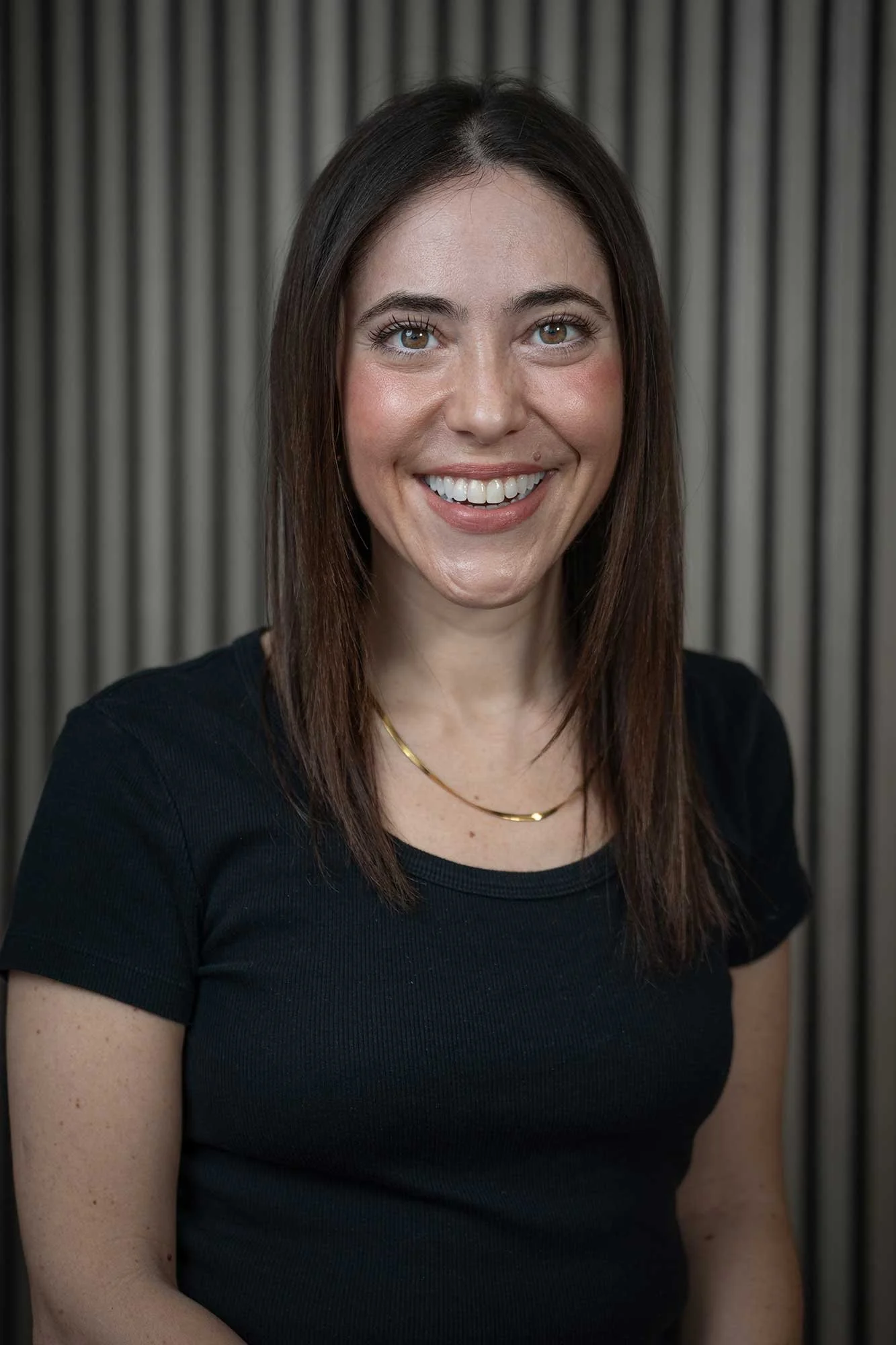 An experienced London therapist with shoulder-length brown hair, smiling, wearing a black top and a gold necklace, standing in front of a vertical striped background.