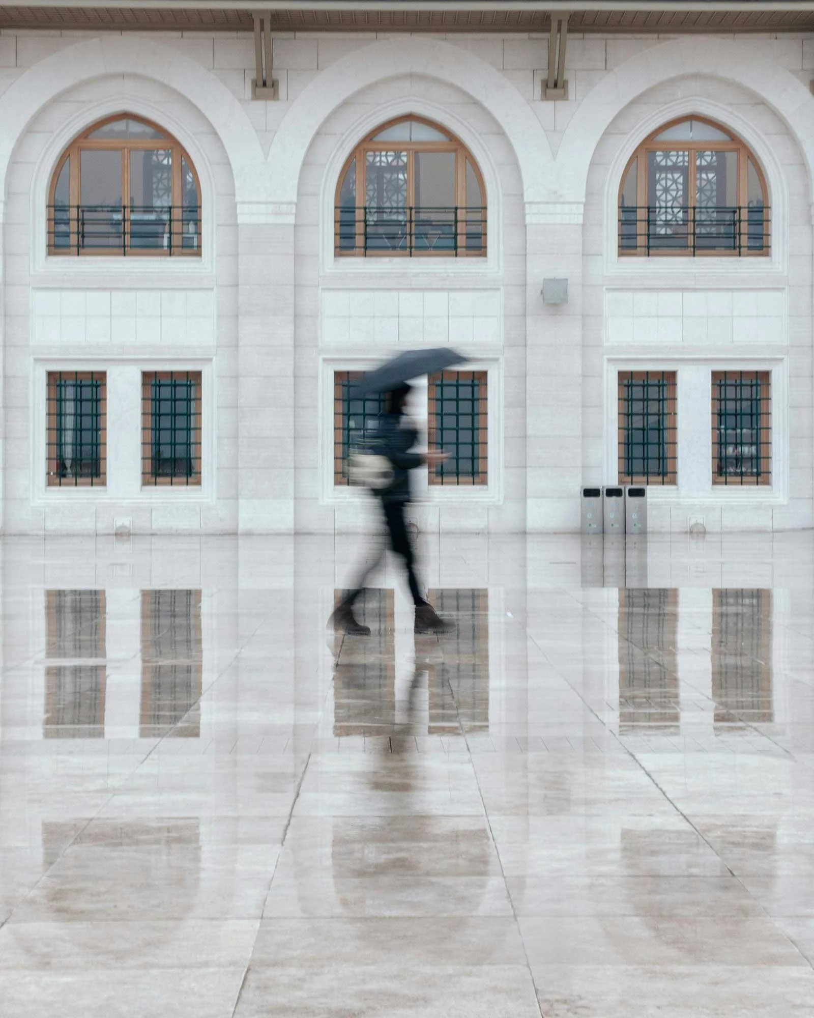 A person wearing dark clothing and holding an umbrella walks through a spacious, shiny hallway with a polished tiled floor and large arched windows.