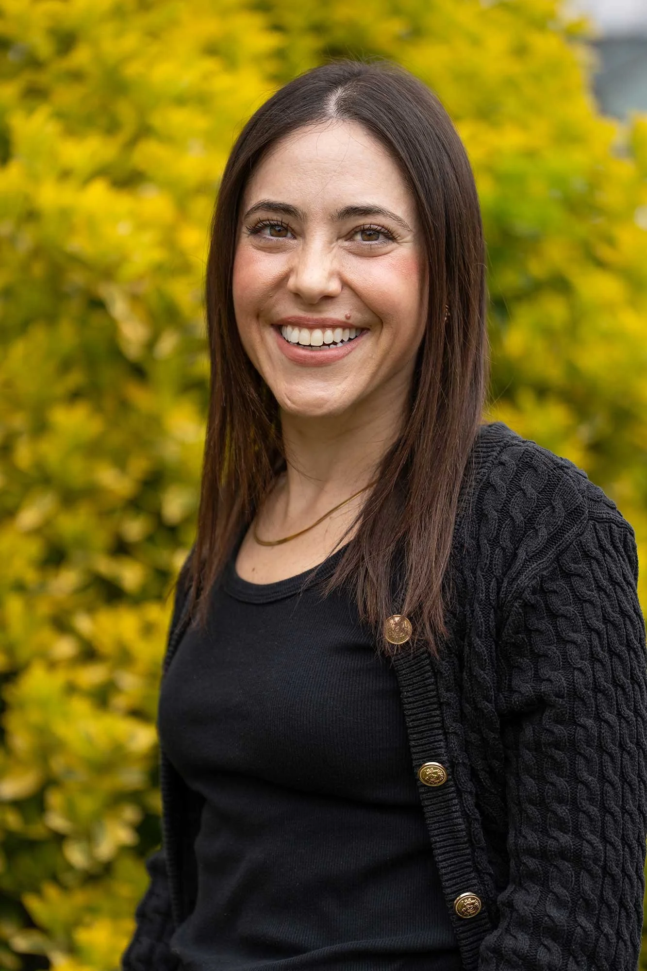 A smiling woman with long brown hair standing outdoors in front of yellow foliage, wearing a black top and a dark cardigan with gold buttons.