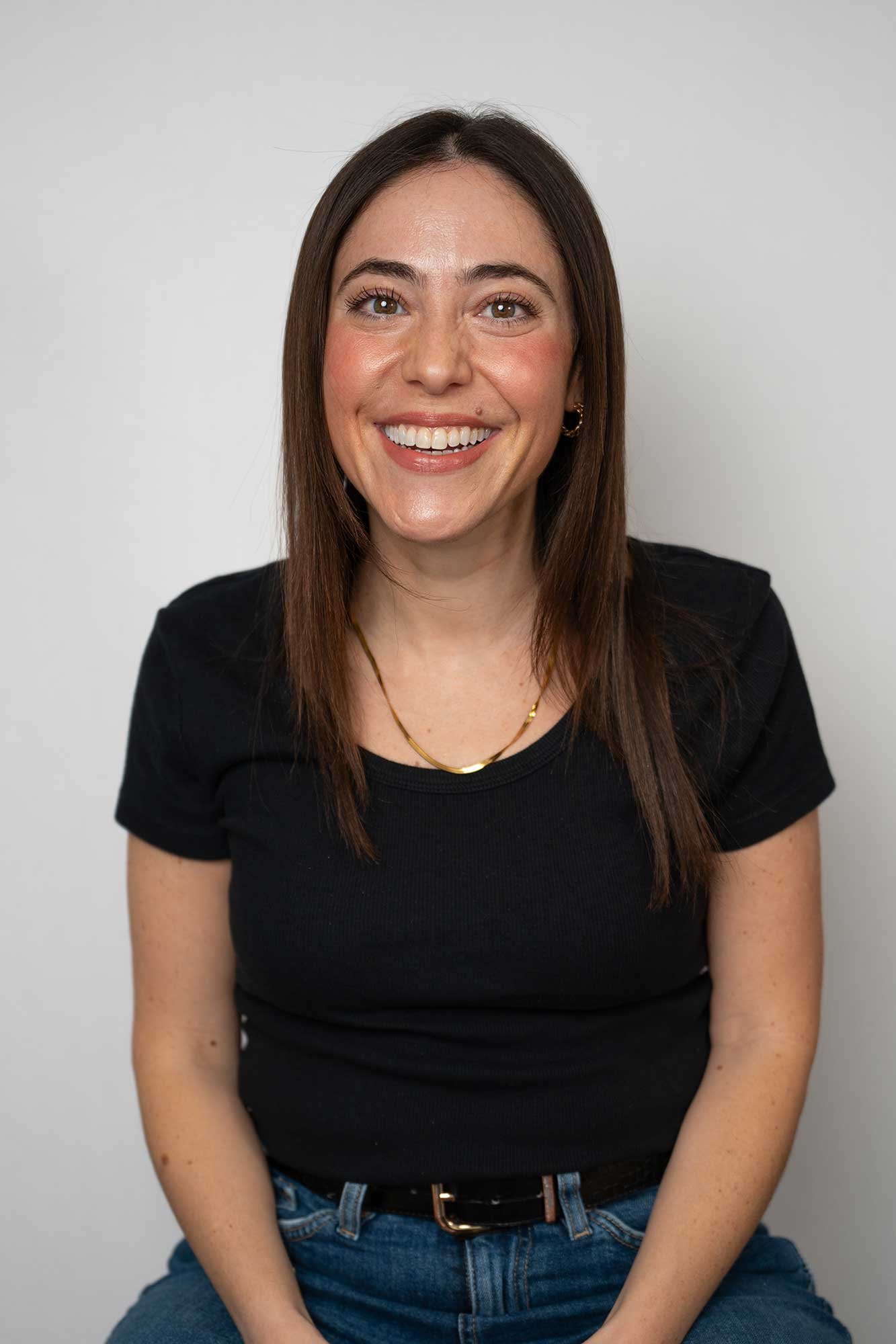 A London Therapist with long brown hair, wearing a black t-shirt and blue jeans, smiling while sitting against a plain light gray background.