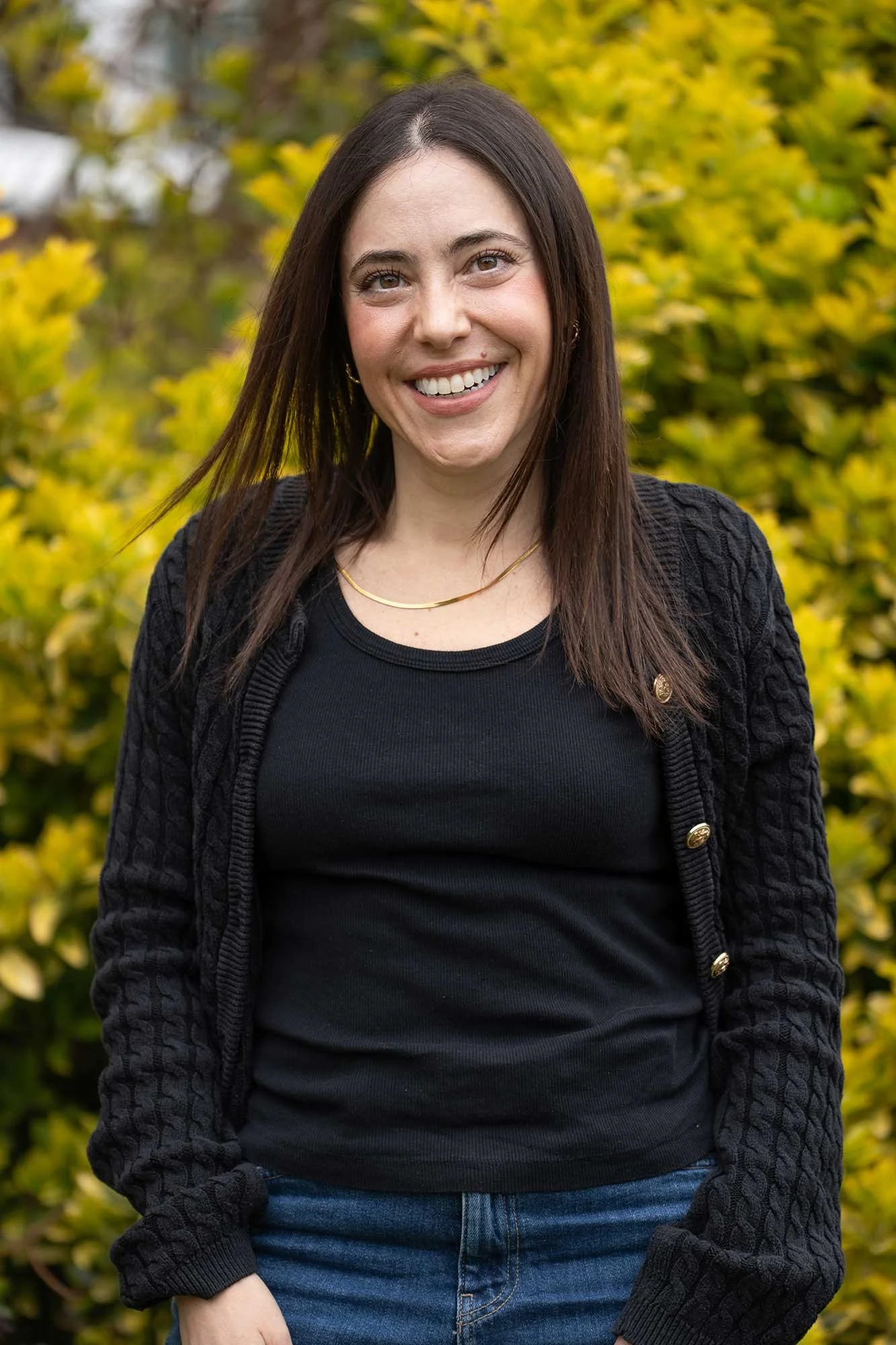 A London Therapist with brown hair wearing a black sweater and denim jeans, standing outdoors in front of yellow leafy bushes, smiling at the camera.