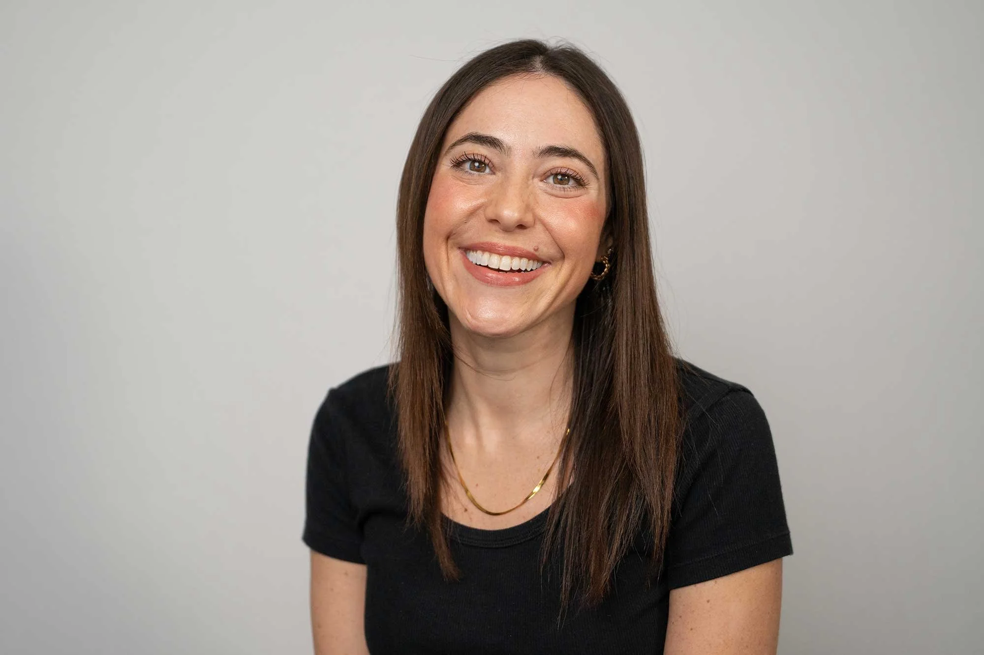 A London Therapist with long brown hair, smiling, wearing a black top, gold jewelry, and earrings, against a plain light grey background.
