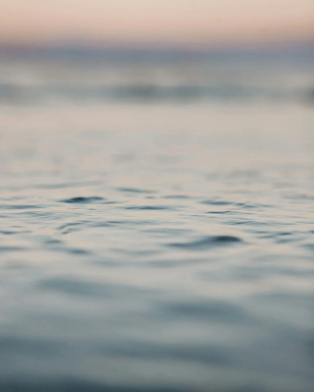 Close-up of calm ocean water with gentle ripples, blurred horizon, and pastel-colored sky at sunset or sunrise.