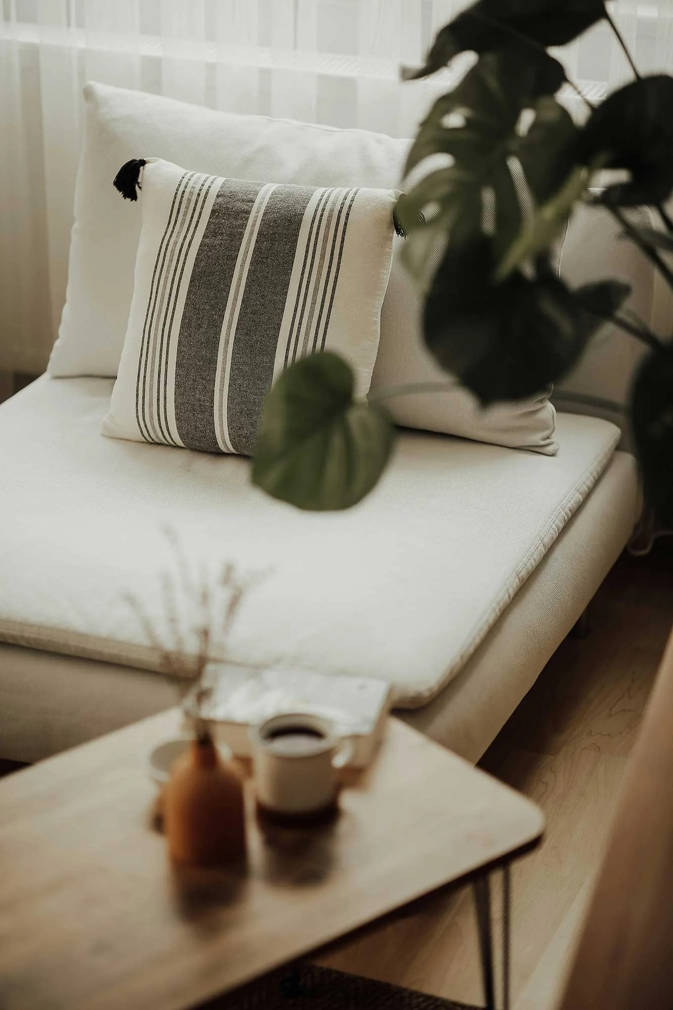 A discreet therapy space in Central London with a white sofa featuring a decorative pillow with gray stripes and tassels, a plant in the foreground, a small wooden table with a brown bottle and a roll of tape, and soft natural lighting.