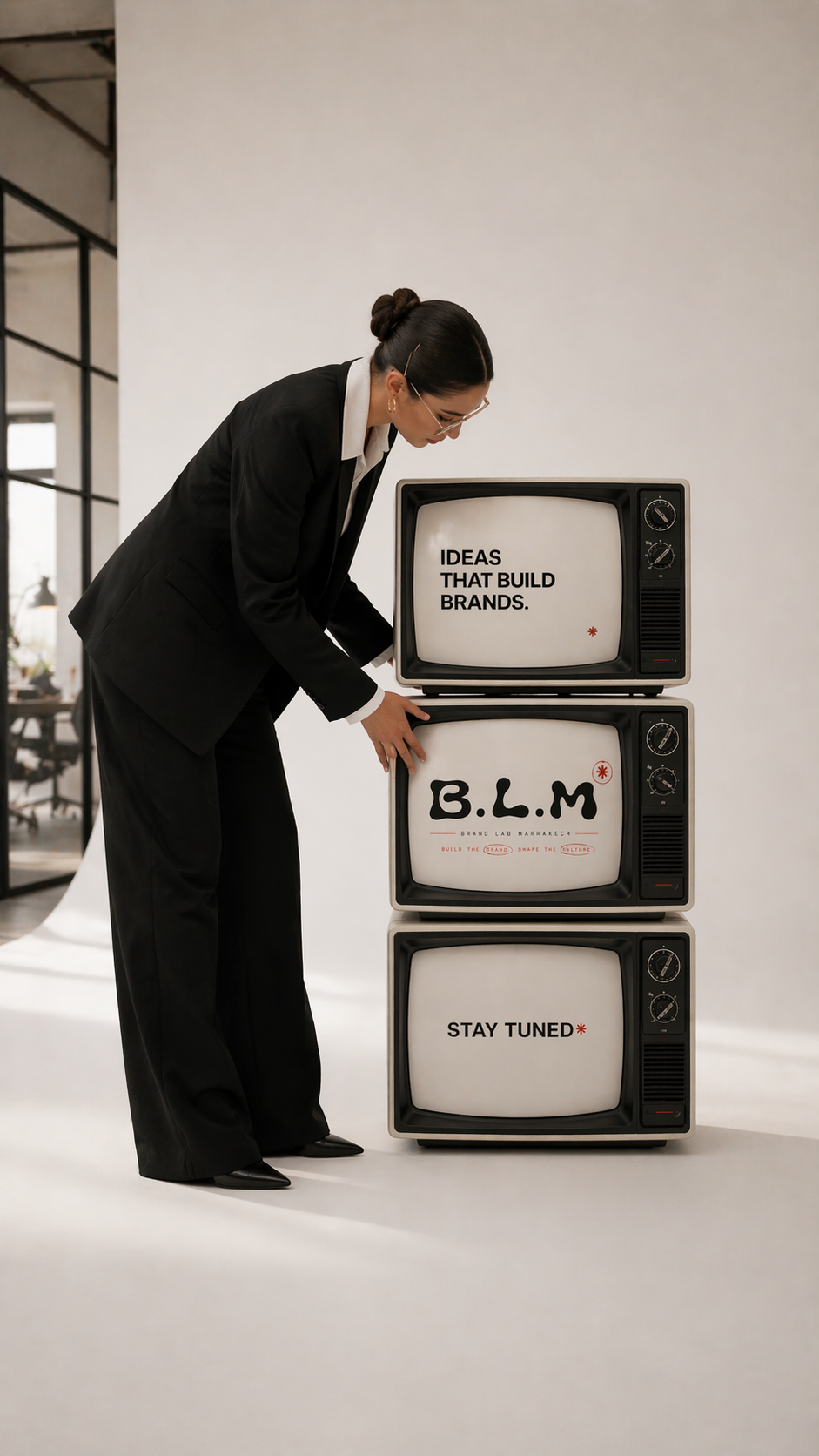 A woman in a black suit is standing beside three stacked vintage televisions. The top TV screen displays the text "IDEAS THAT BUILD BRANDS," the middle TV has "B.L.M" with additional smaller text, and the bottom TV reads "STAY TUNED."