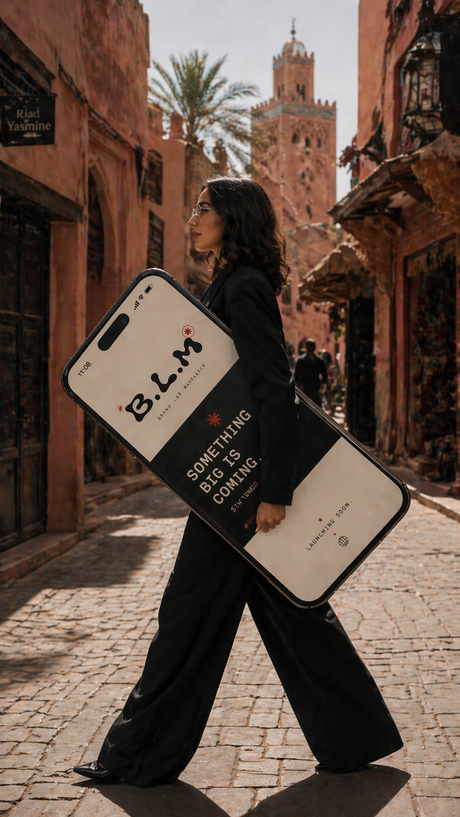 A woman walking down a cobblestone street in Morocco, carrying a large phone-shaped sign promoting an upcoming launch, with a reddish tower and palm trees in the background.