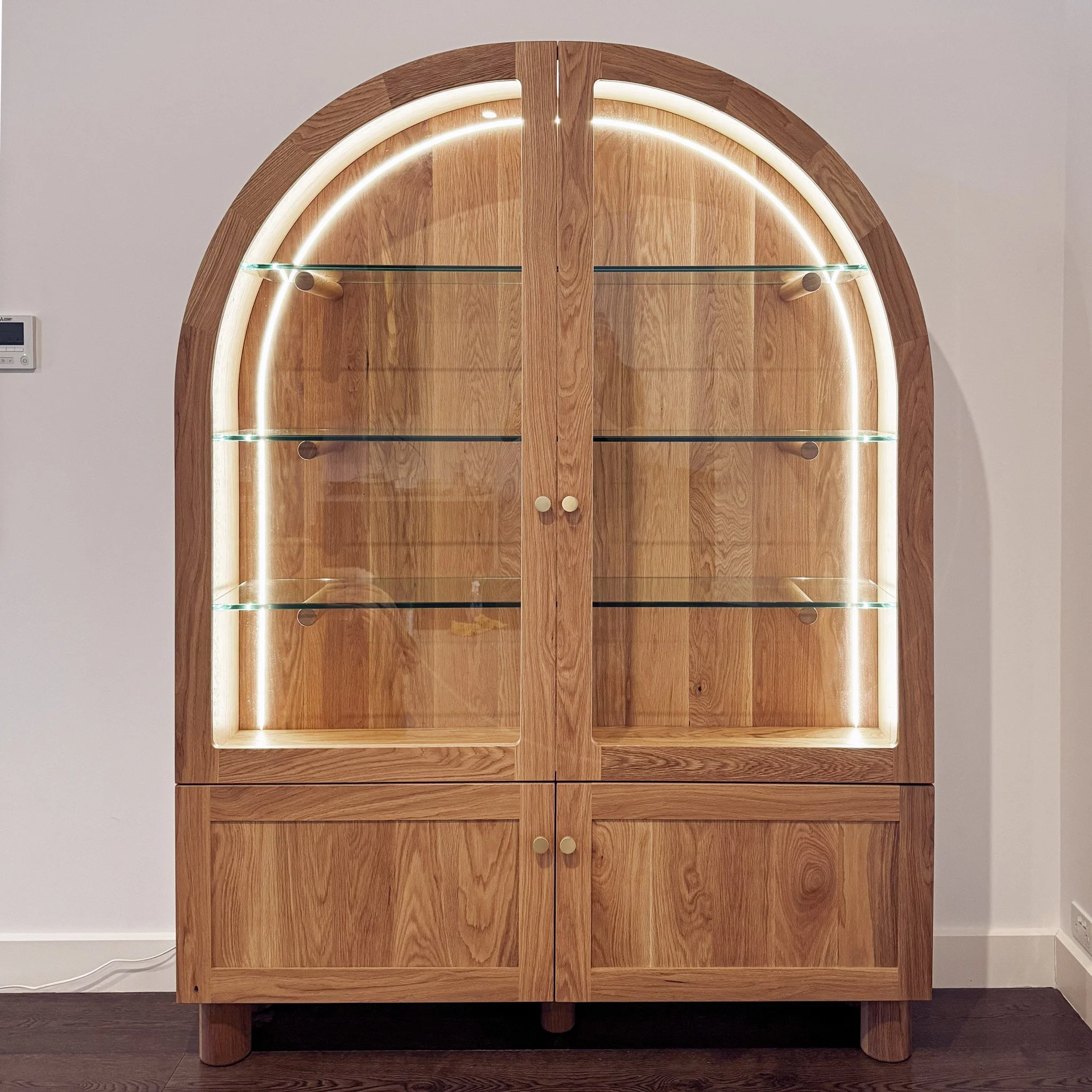 A wooden cabinet with an arched top, two glass shelves, and small round knobs on doors, illuminated by embedded LED lights, set against a plain wall.