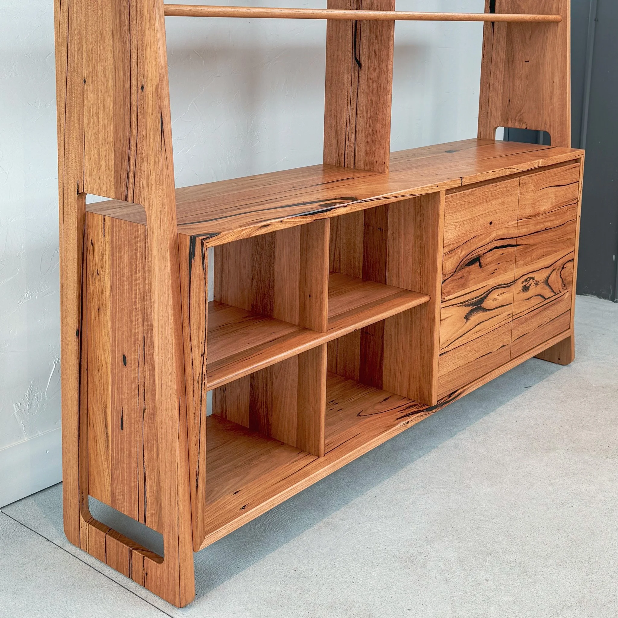 A wooden bookshelf with multiple compartments, made from reddish-brown wood with a natural grain pattern, positioned against a light-colored textured wall and resting on a gray carpeted floor.