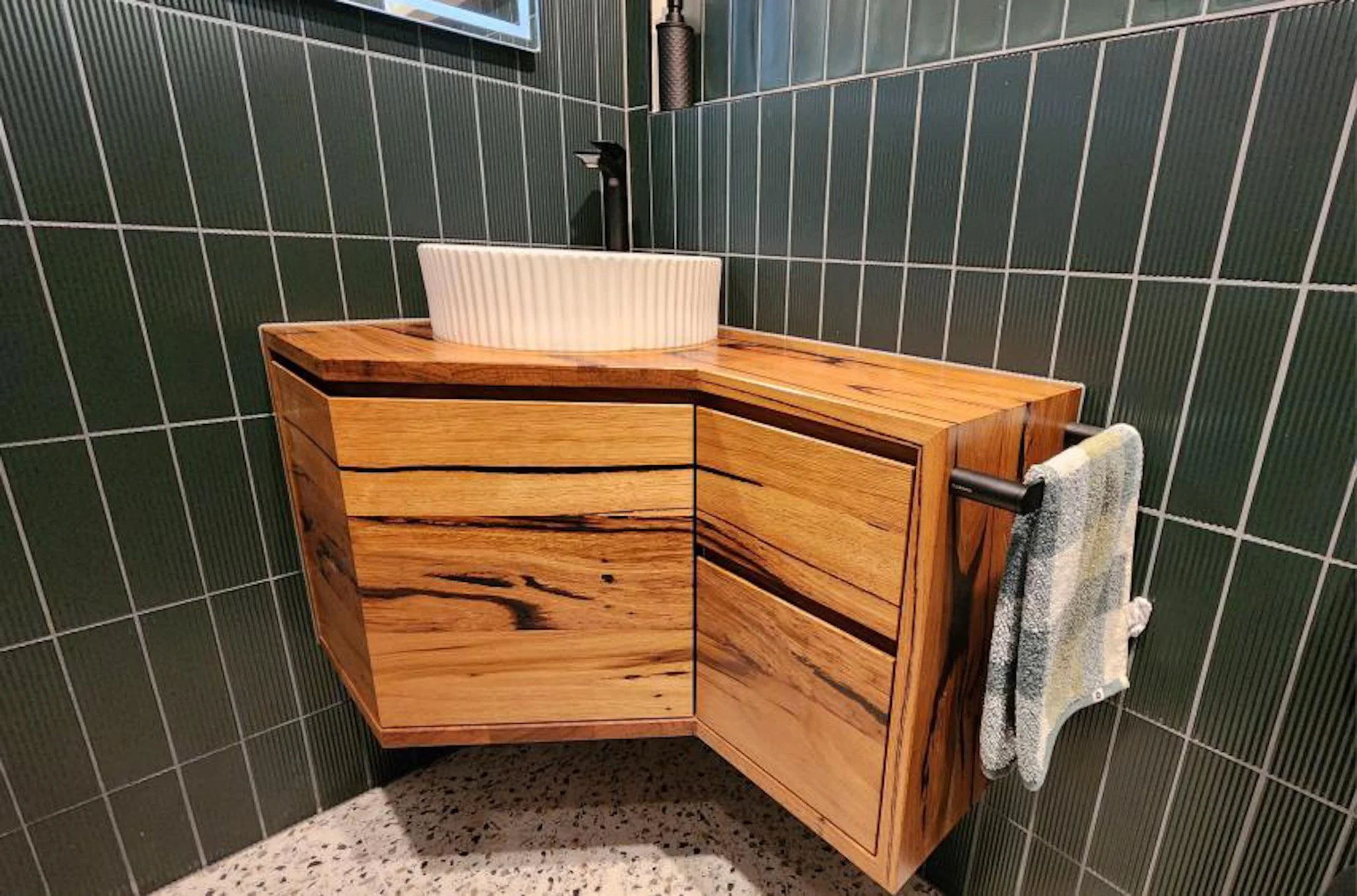 A wooden bathroom vanity with a custom round white vessel sink, black faucet, and a textured hand towel hanging from a rail on the right side. The background features dark green tiled walls and speckled beige and black tile flooring.