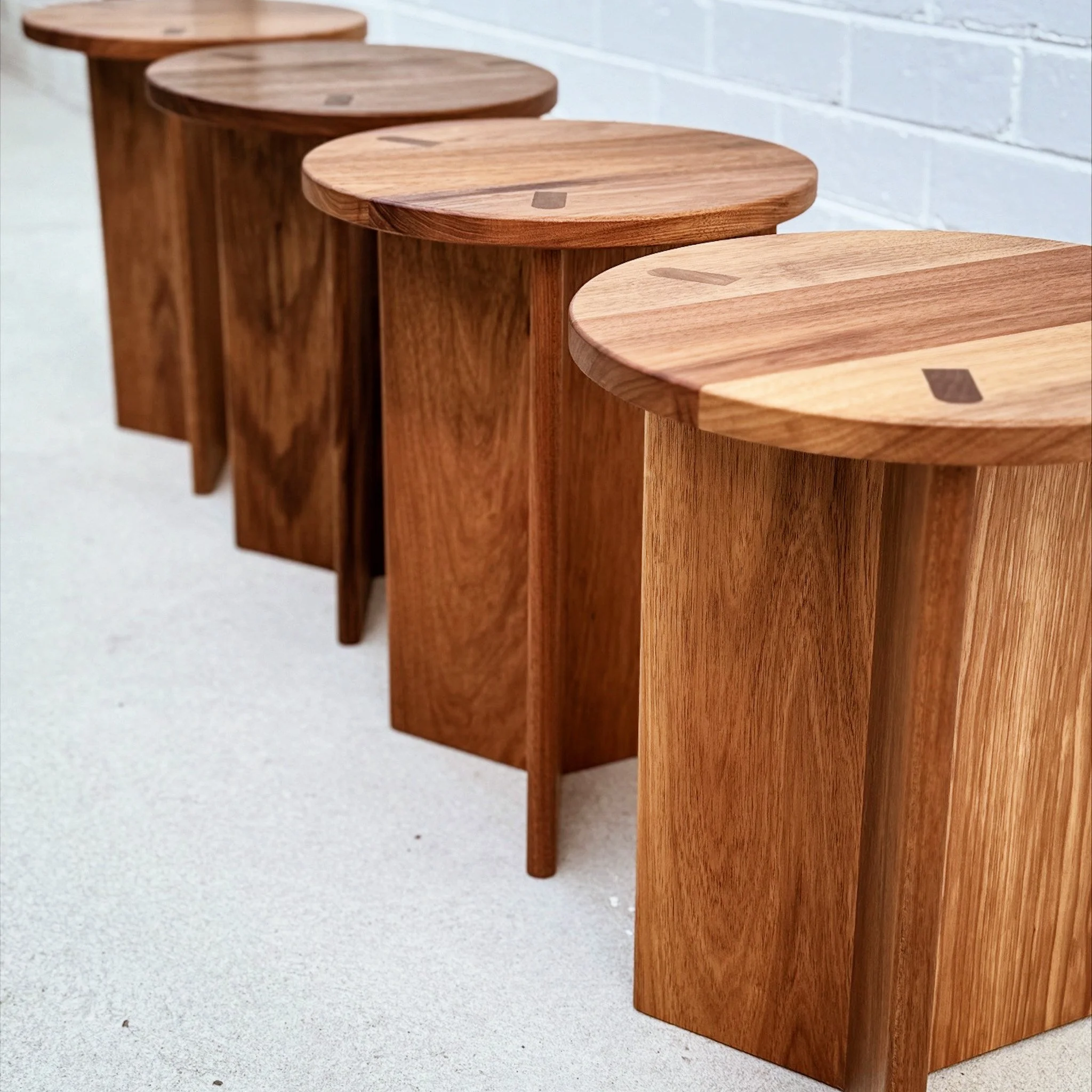 A row of four wooden stools with round seats and four legs each, placed along a white brick wall on a light-colored floor.