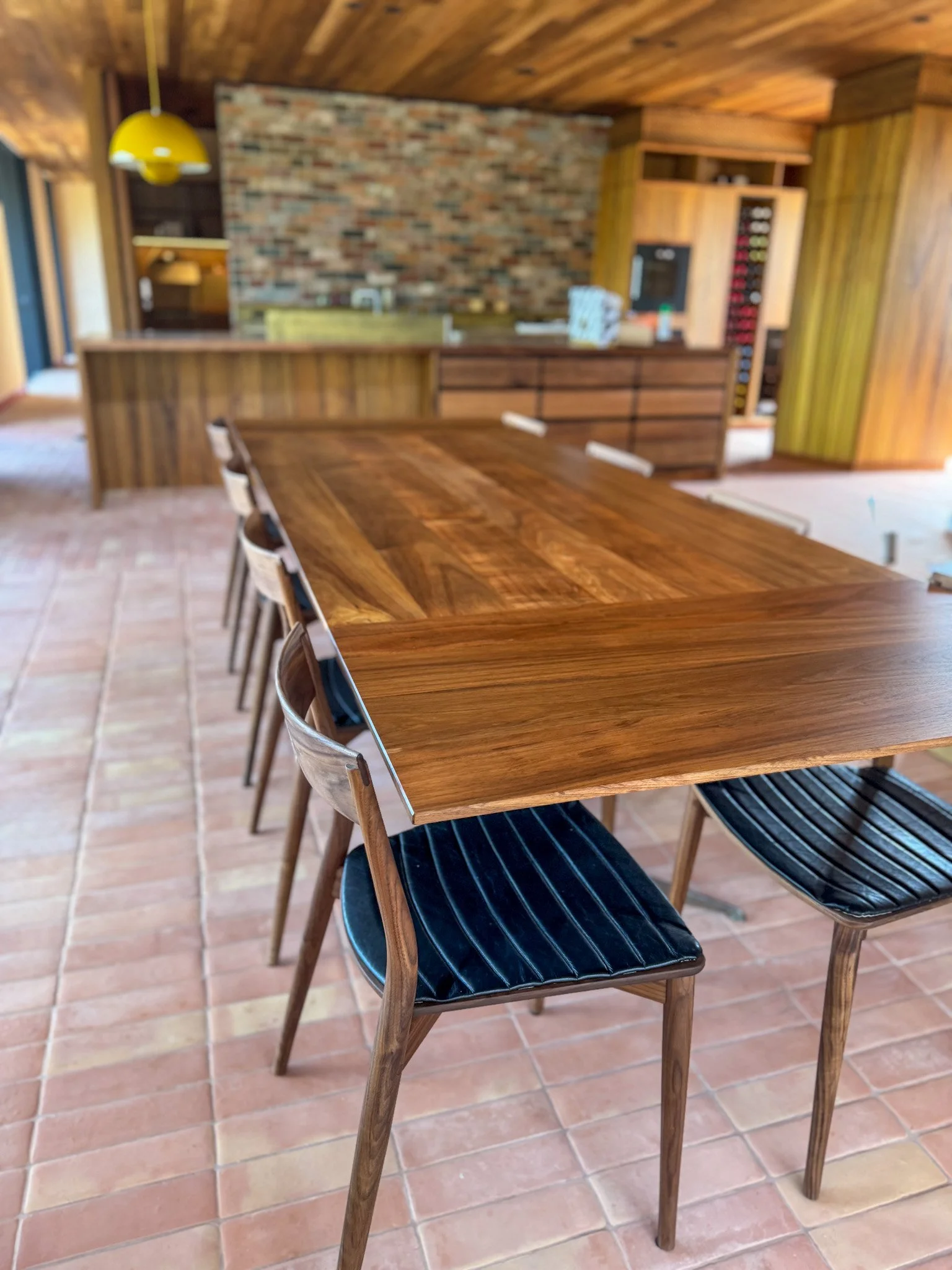 A wooden dining table with black cushioned chairs with wooden frames in a rustic kitchen with a brick backsplash and wooden cabinets.