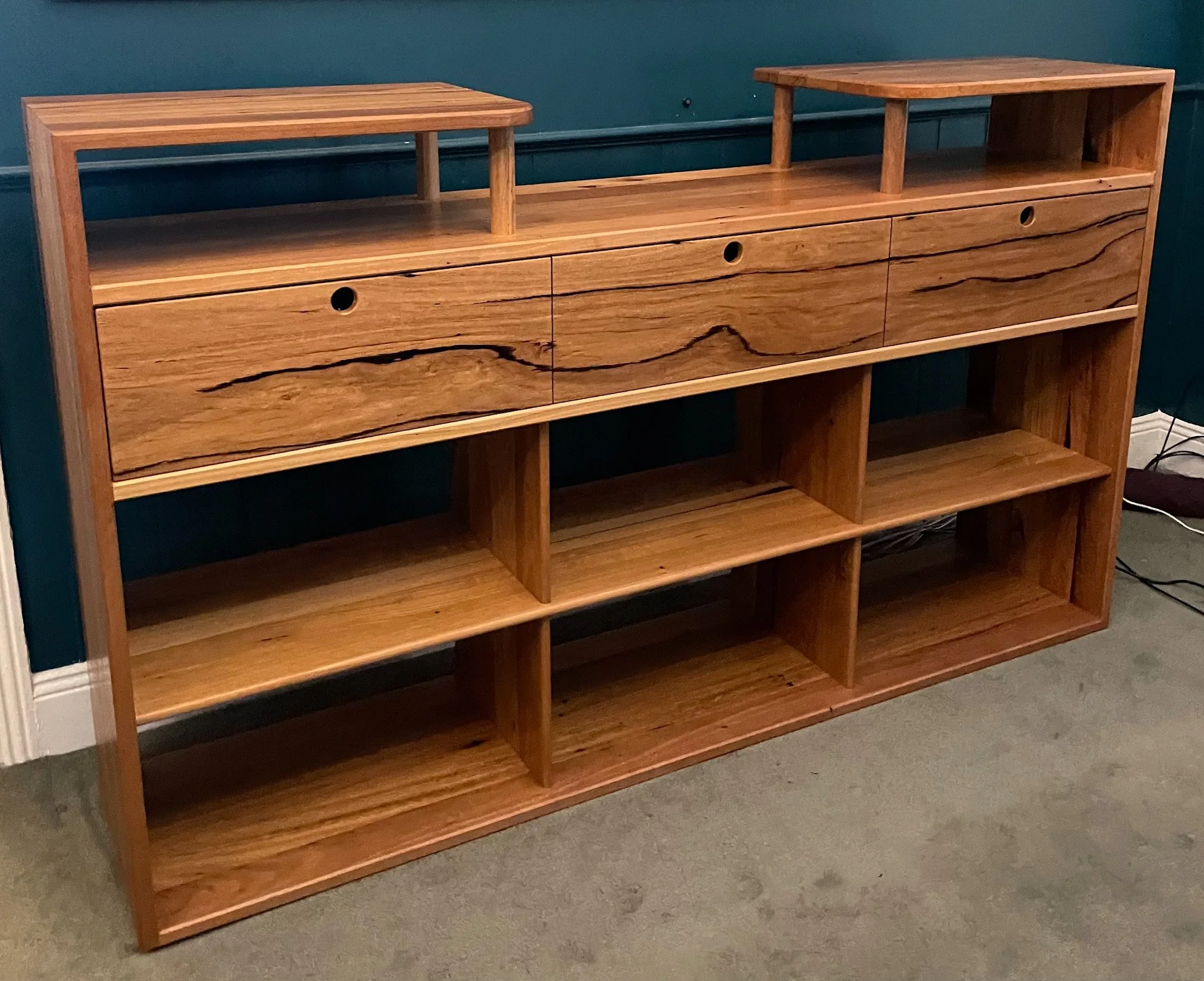A timber DJ console, hifi record play cabinet, bookshelf with multiple open shelves for vinyl and two small drawers at the bottom, set against a teal wall with electrical cords visible beside it.