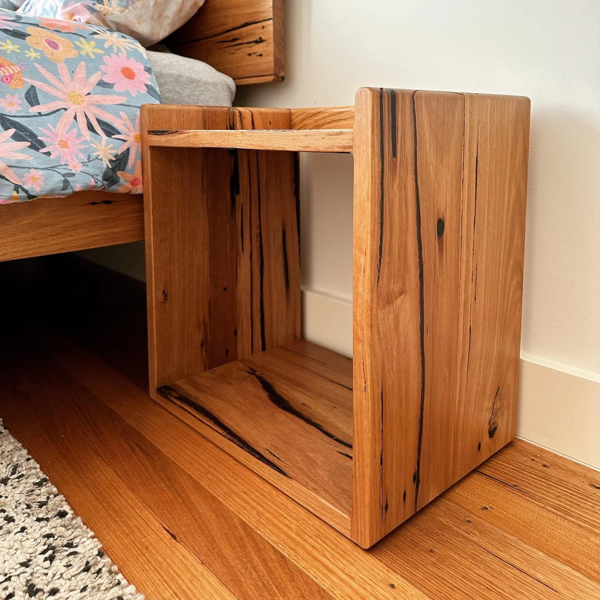 Close-up of a wooden nightstand with a natural, rustic finish, next to a bed with floral bedding and a gray pillow.