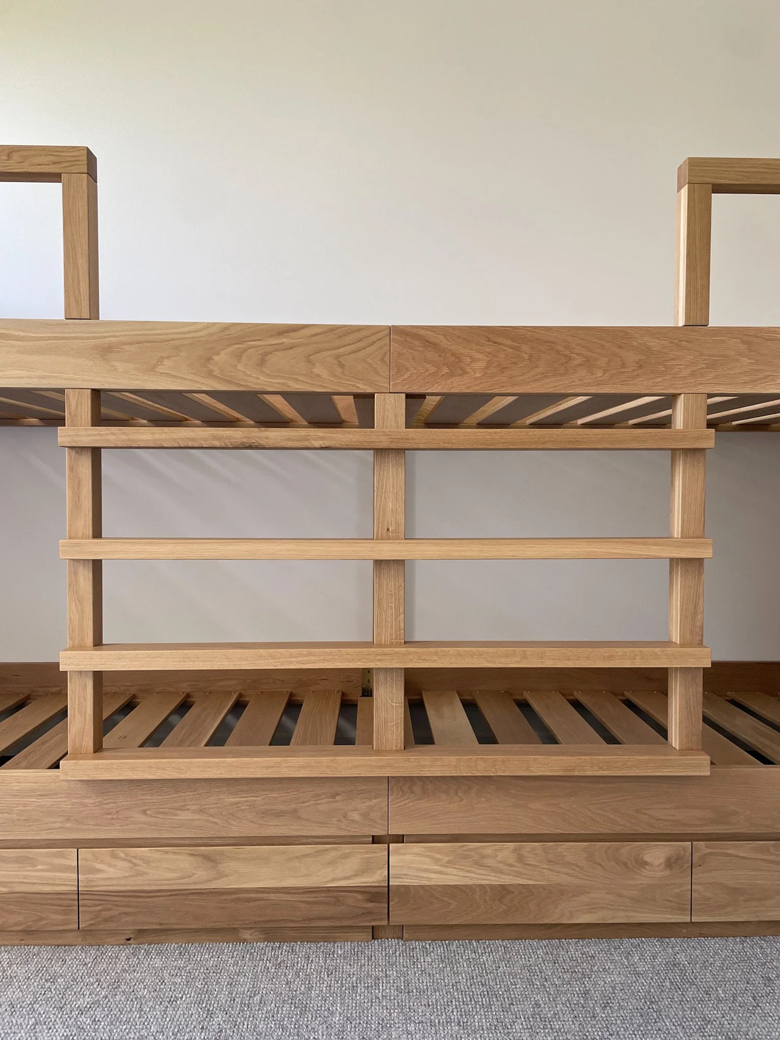 A light oak bunk bed frame with slatted base and matching headboard, positioned against a plain wall with a gray carpeted floor.