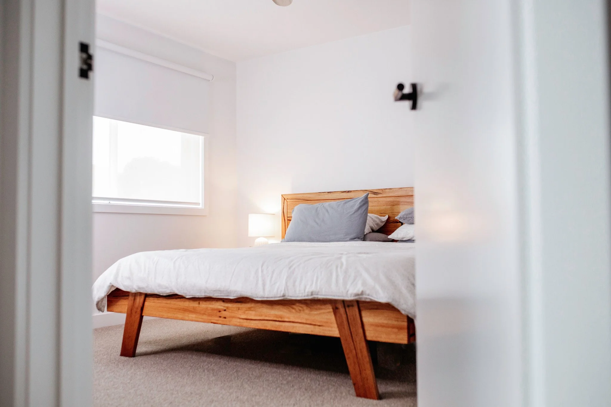 timber bed with a wooden frame, gray pillows, and a lamp on a bedside table in a bright, minimalist bedroom with white walls and a large window.