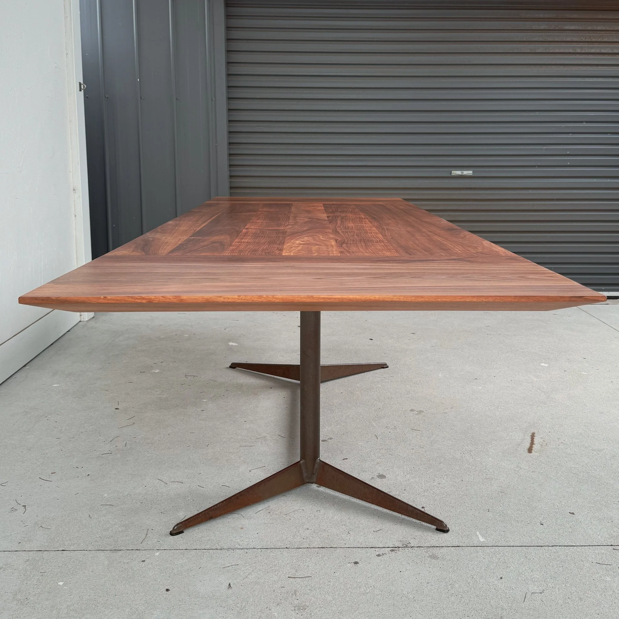 A wooden table with a metal base in front of a gray metal wall and a white wall.