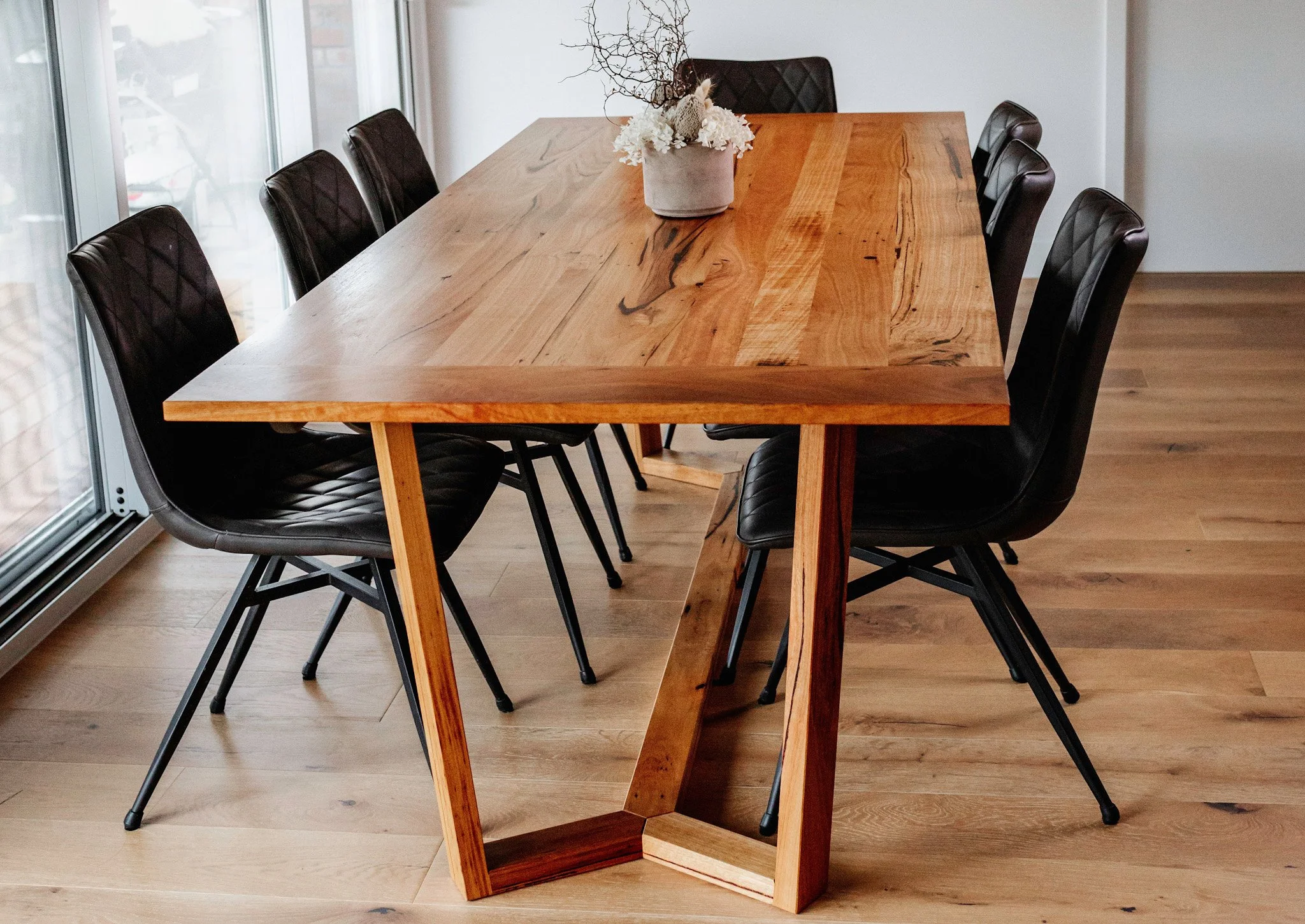 A wooden dining table with a vase of white flowers and decorative branches in the center, surrounded by six black leather chairs, near large windows in a modern home.