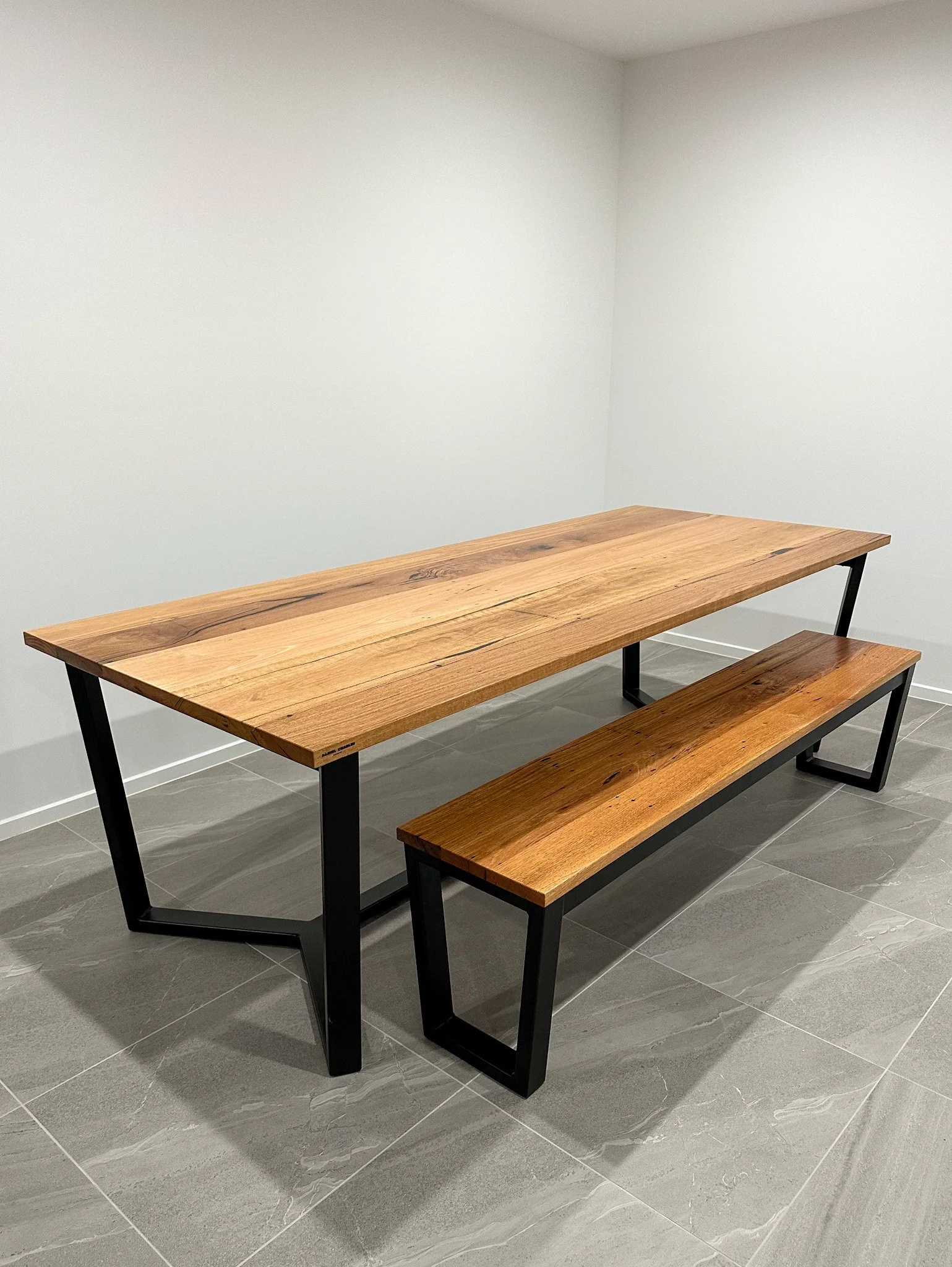 Wooden dining table with matching bench and black metal frame, set on gray tiled floor in a room with plain white walls.