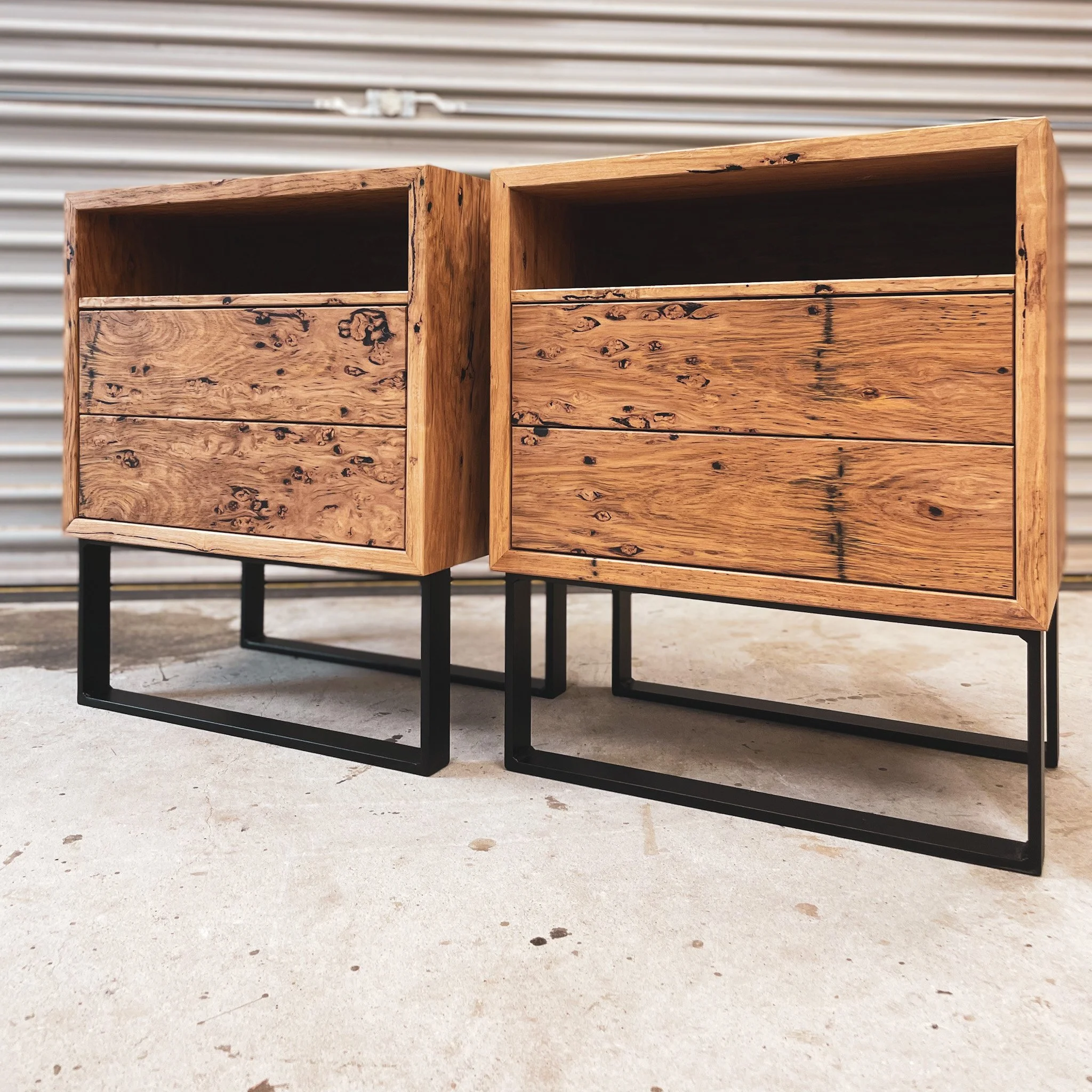 Two wooden cabinets with black metal bases, placed outdoors on a concrete surface, against a backdrop of a white corrugated metal wall.