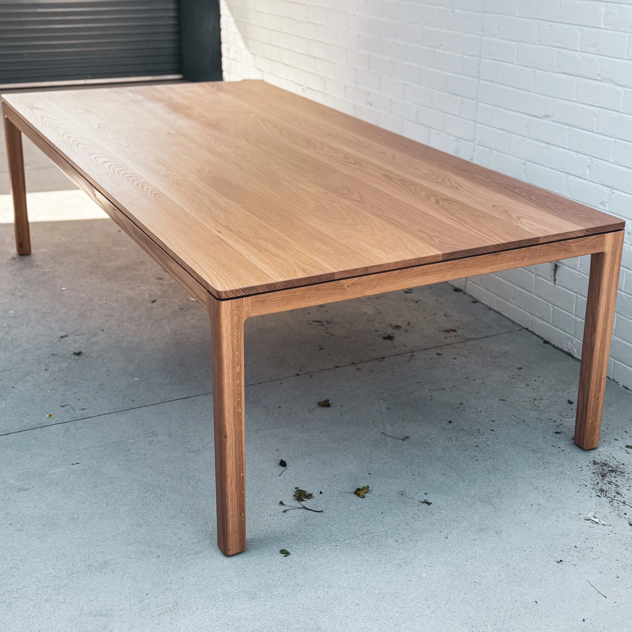 A wooden table with a smooth top and four legs, placed outdoors against a white brick wall, with a concrete ground and some fallen leaves underneath.
