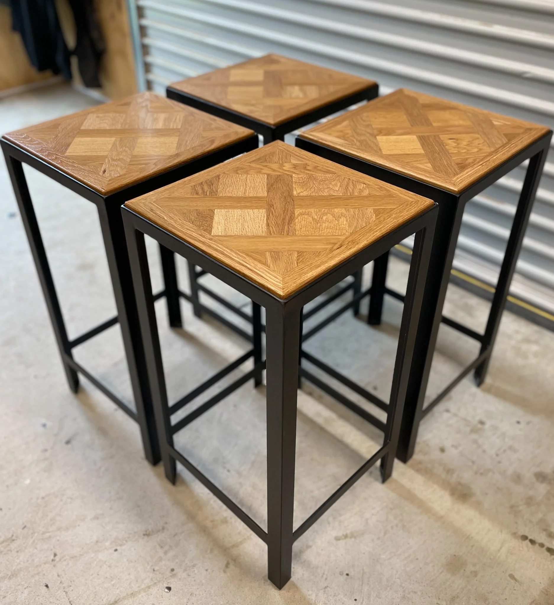 Four small stools with wooden tops featuring geometric parquet patterns and black metal legs, placed on a concrete floor in front of a metal roll-up door.