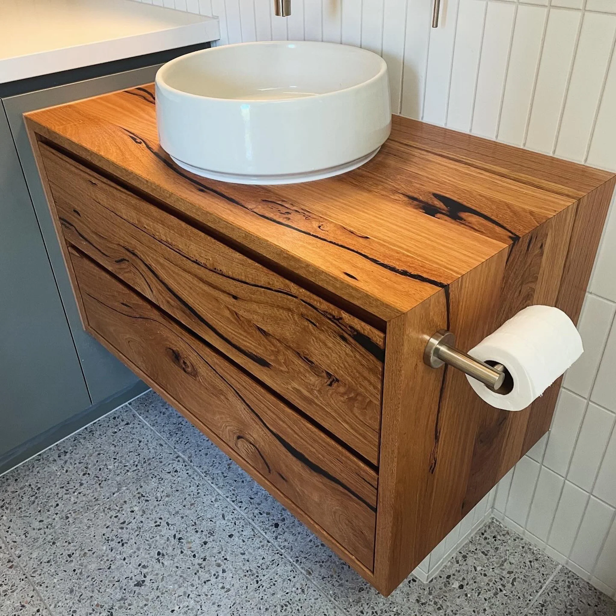 A white ceramic bowl is placed on a wooden counter next to a toilet paper roll holder in a bathroom. The wall behind has white vertical paneling, and the floor is covered with speckled terrazzo tiles.