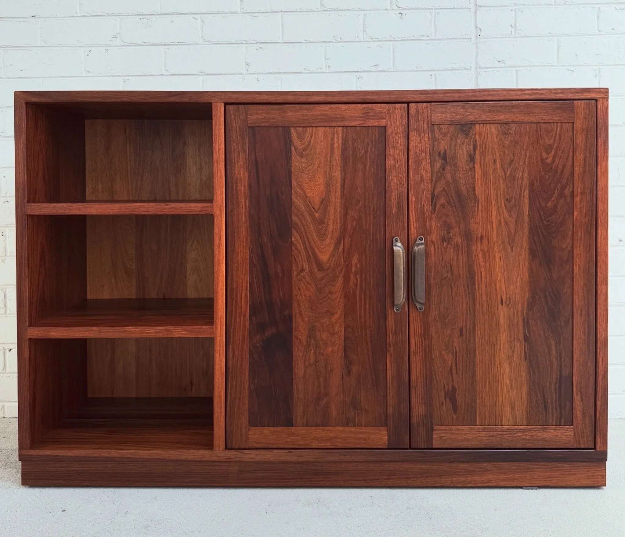 A wooden cabinet with a closed door, two handles, and open shelves against a white brick wall.