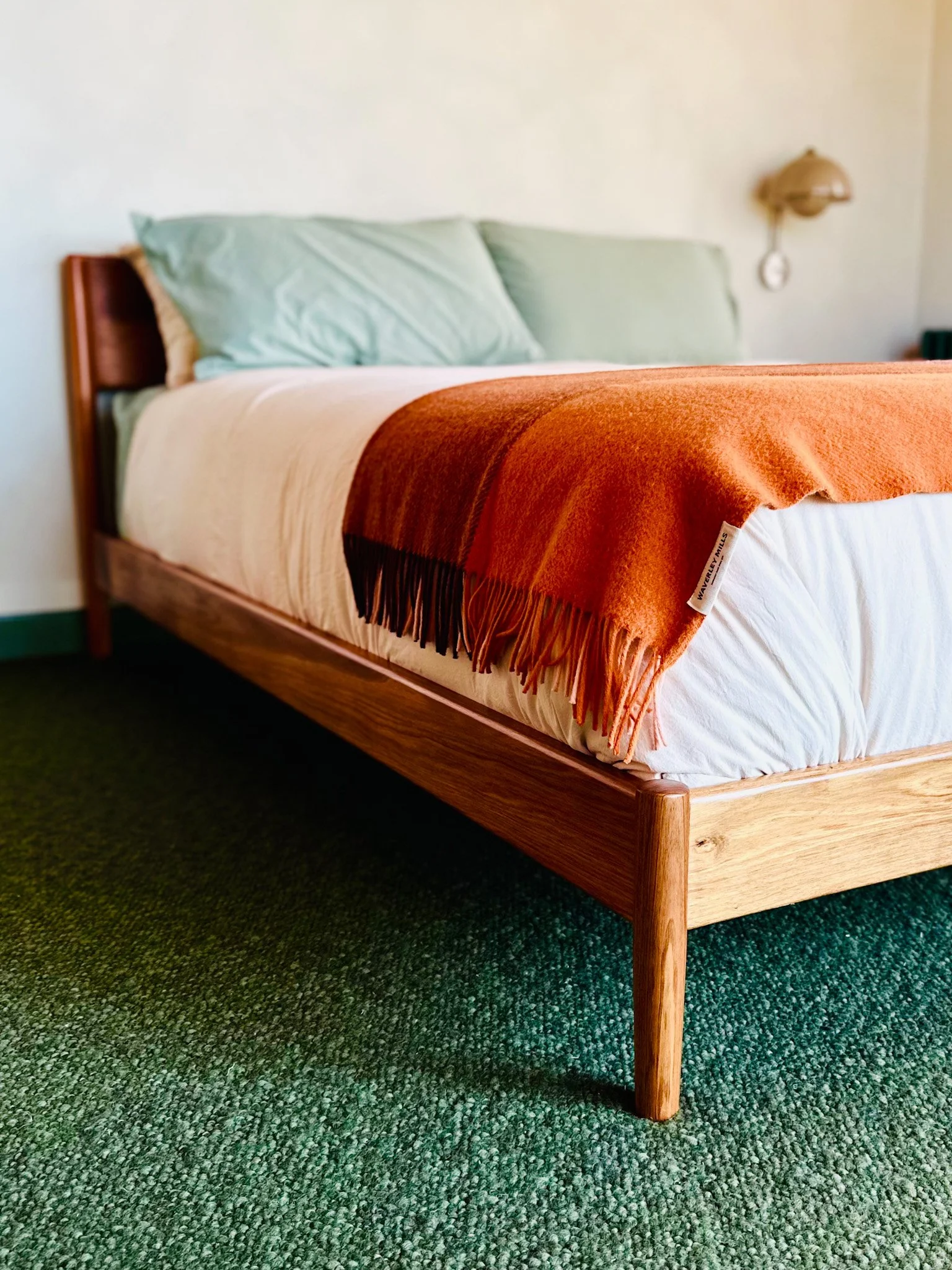 A bed with green pillows, a white blanket, and an orange blanket, in a room with green carpet and a wall-mounted lamp.