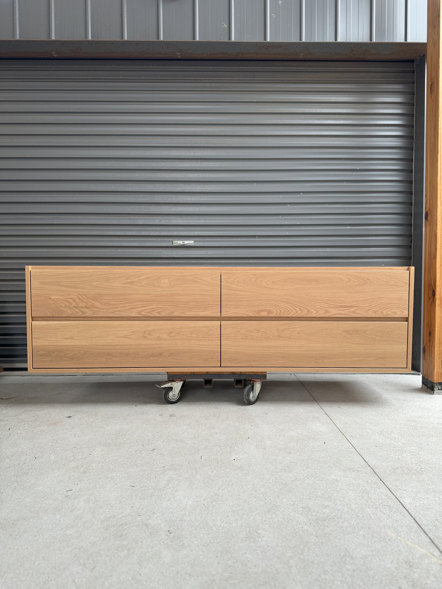 A wooden cabinet on wheels is positioned in front of a gray metal roll-up door and a wooden post inside a warehouse or garage.