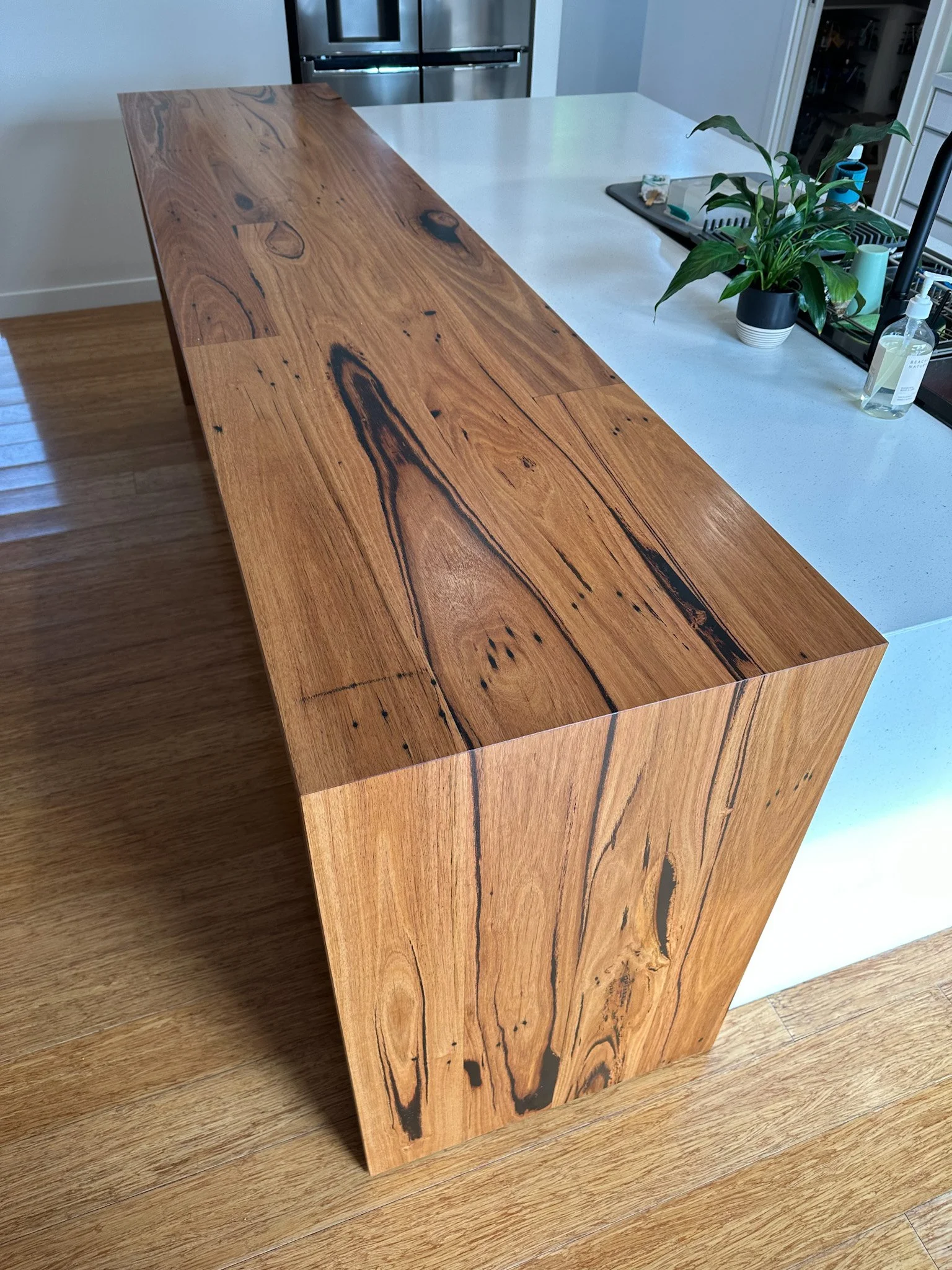 A wooden kitchen island with a natural finish in front of a white countertop, a potted plant, a bottle of hand sanitizer, and a black dish drying rack visible on the counter.