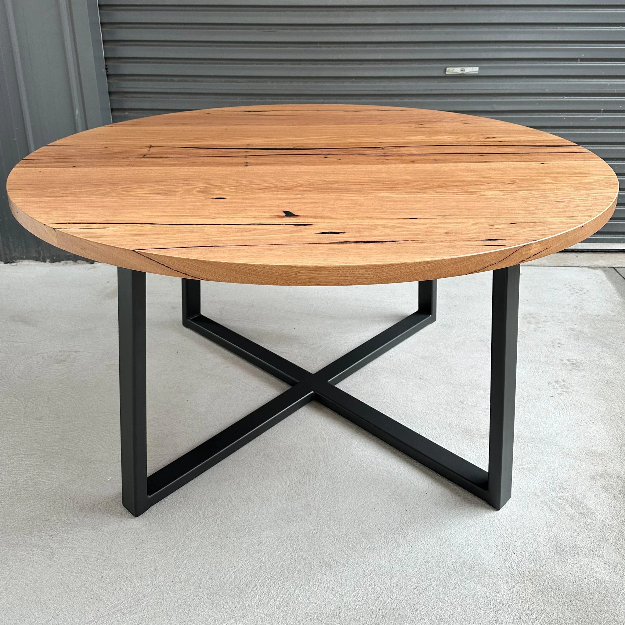 Round wooden table with a metal frame, set against a gray metal roll-up door background, with concrete floor.