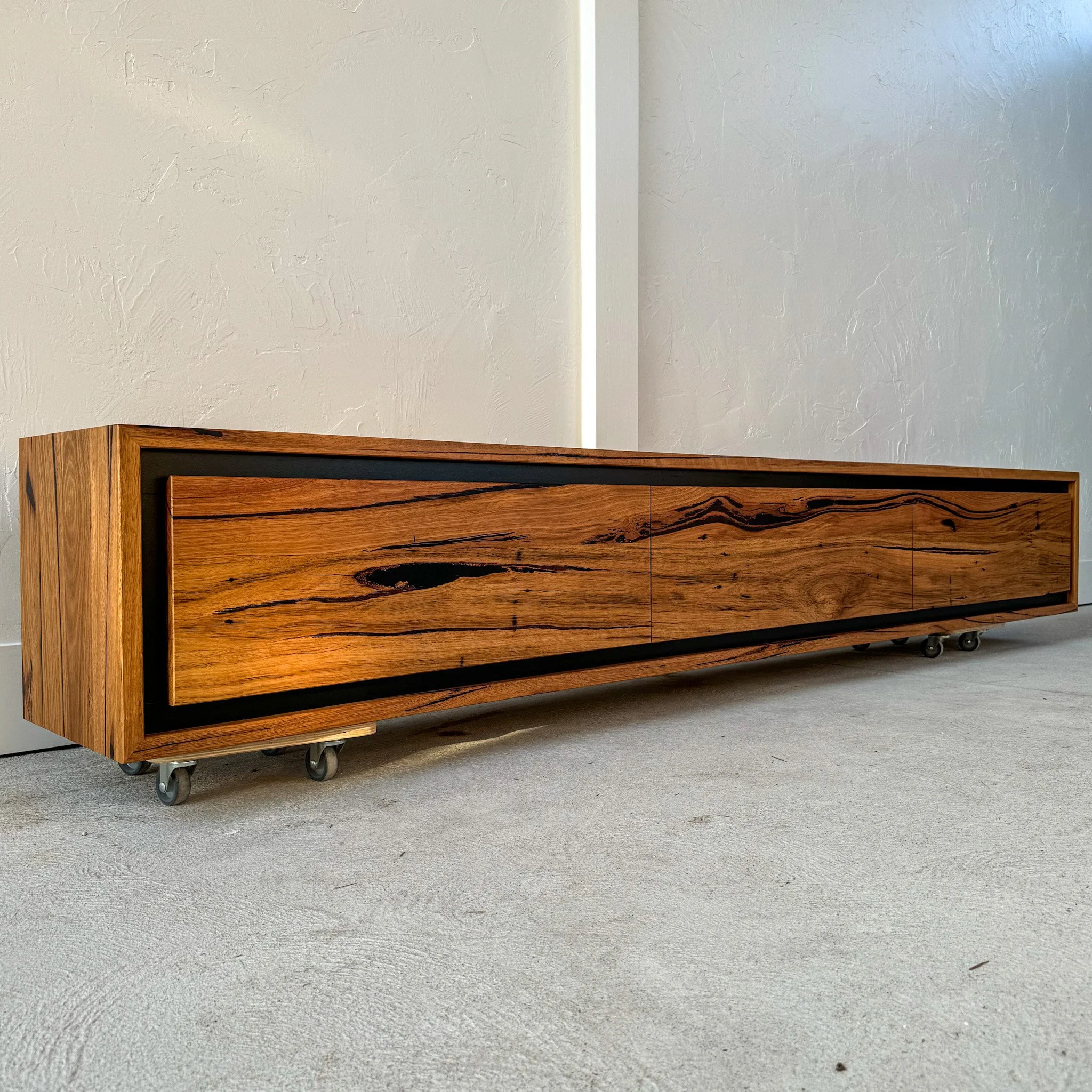 A timber tv cabinet against a textured wall. The cabinet has a natural wood finish with dark streaks and black shadow line trim.