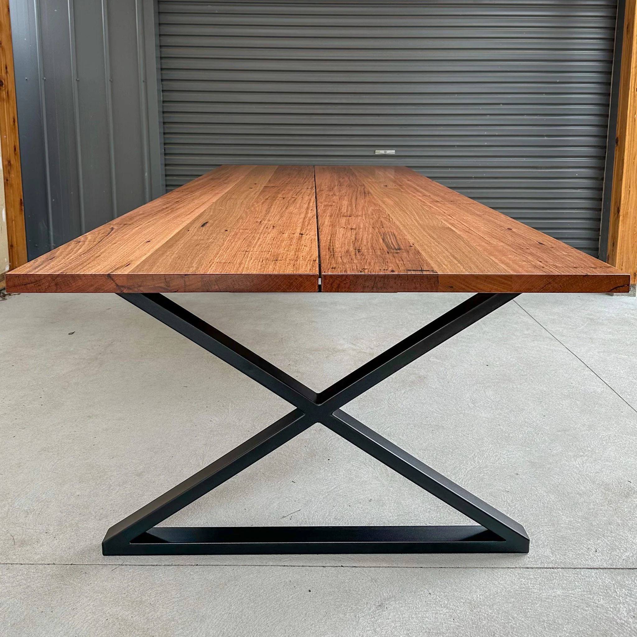 A wooden-top table with a black metal X-shaped base in front of a gray metal roller door, framed by wooden beams.