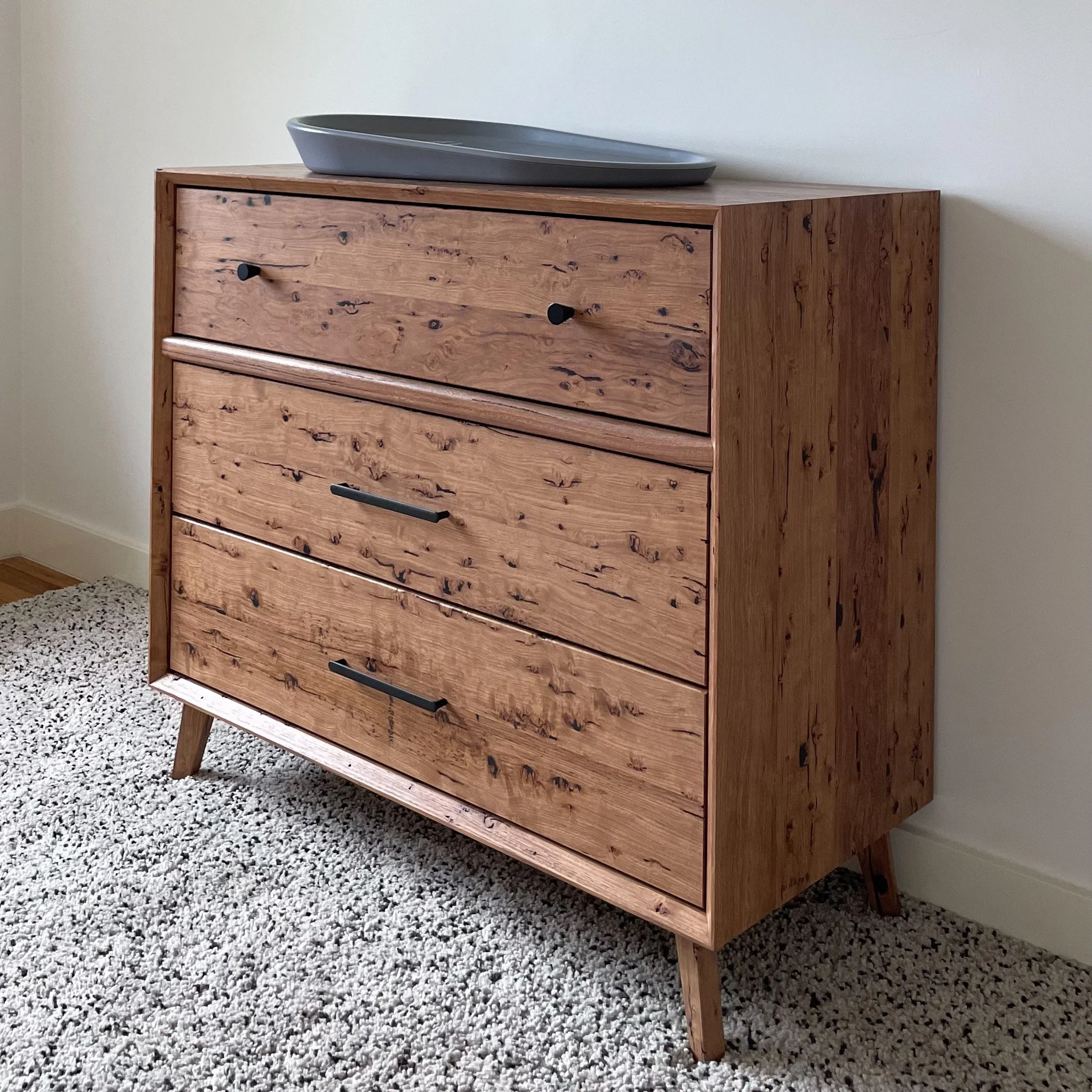 A wooden dresser with three drawers, black knobs, angled legs, and a gray tray on top, positioned against a light-colored wall, with a textured white carpet on the floor.