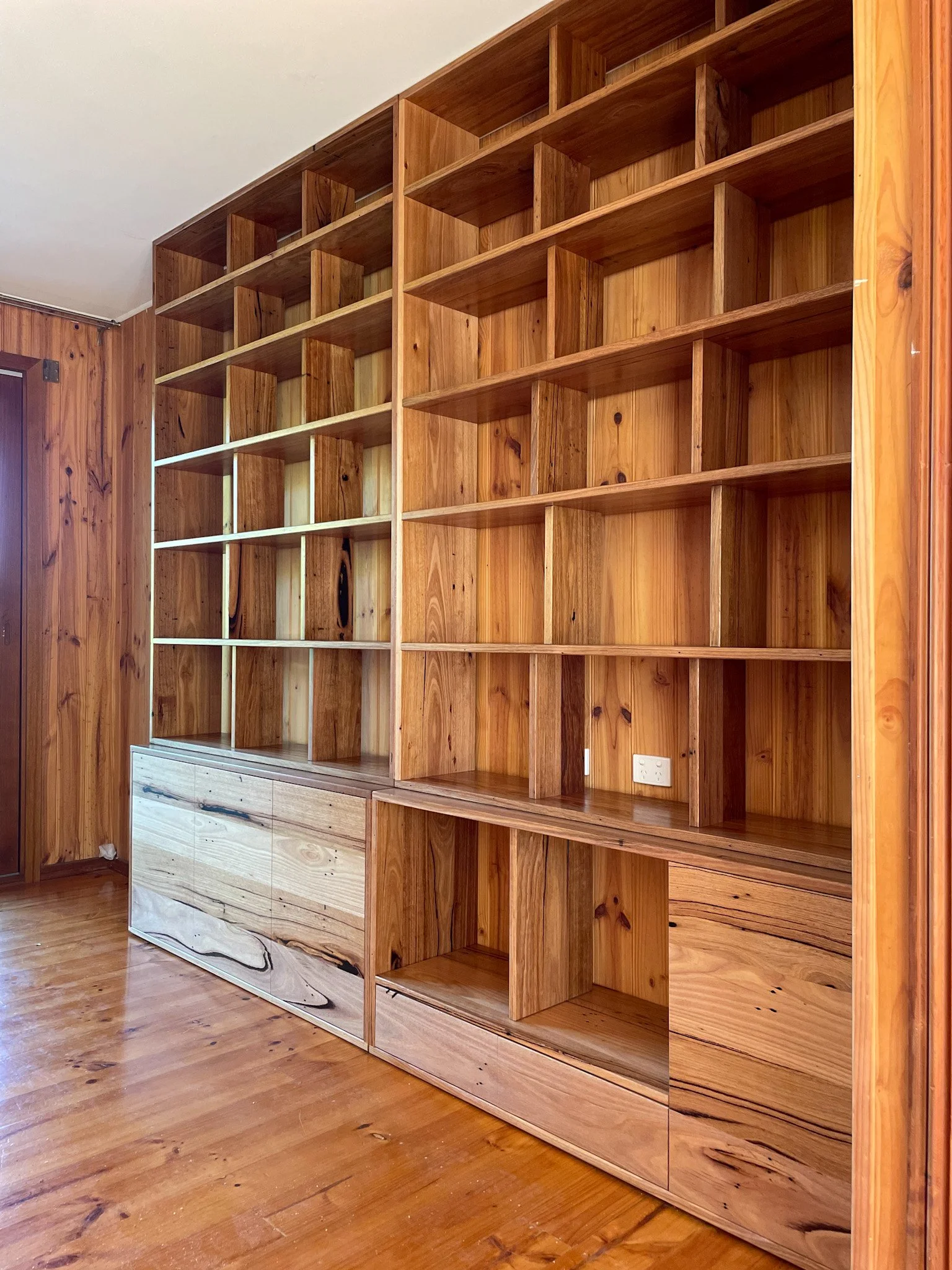 Empty wooden shelving unit with multiple compartments, located in a room with wooden floor and paneled walls.