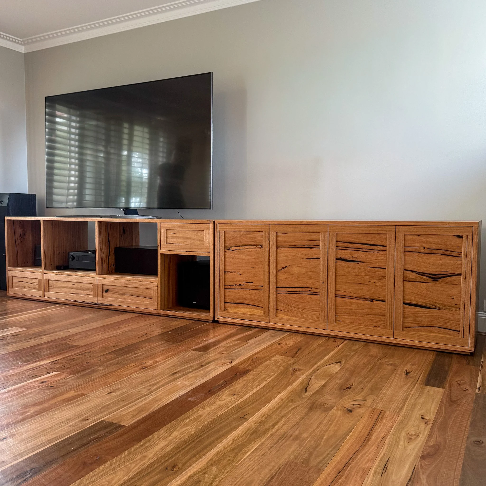 A living room with a large flat-screen TV mounted on a light-colored wall and a wooden entertainment stand below it, featuring multiple open shelves and some electronic devices on the floor to the left. The room has wooden floors.