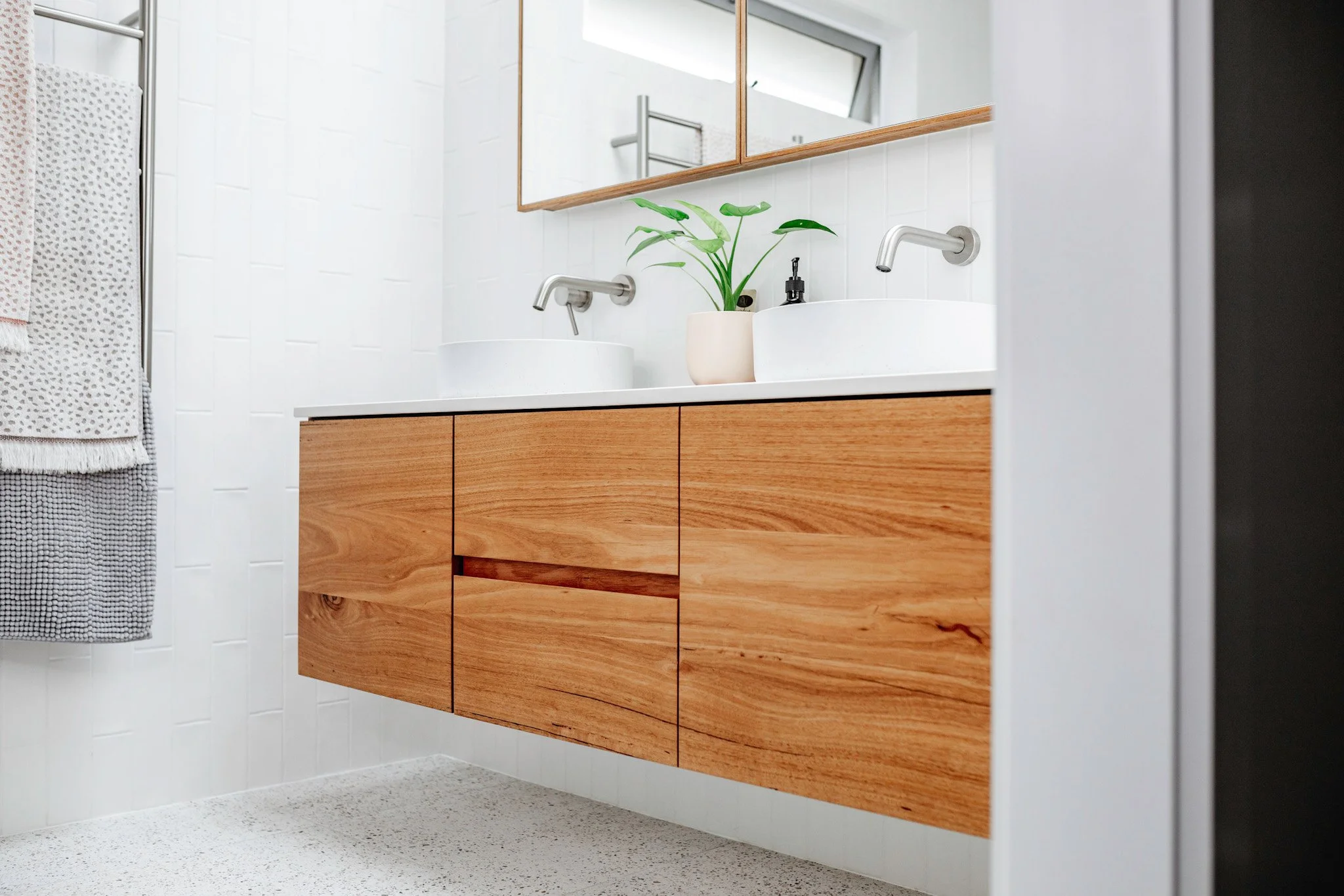 A modern bathroom vanity with a white countertop, two vessel sinks, and a wooden cabinet with drawers. There are two wall-mounted faucets, a green plant in a beige pot, and a large mirror above. To the left, a towel with a dotted pattern hangs on a t