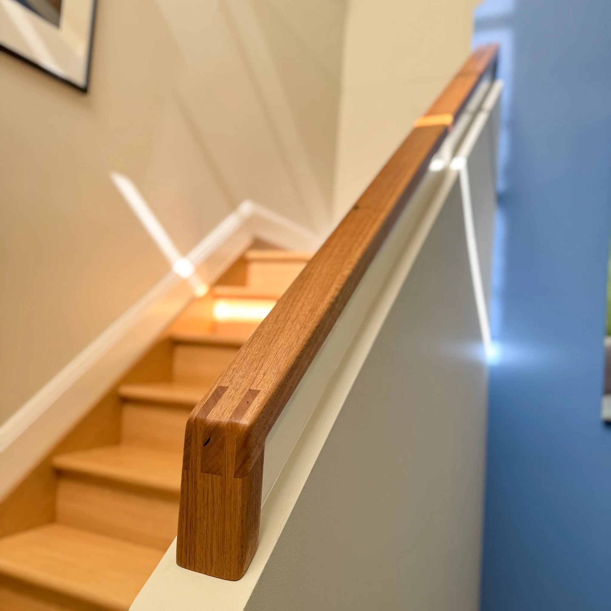 Close-up of a wooden staircase handrail along beige wall with picture frame visible in the background.