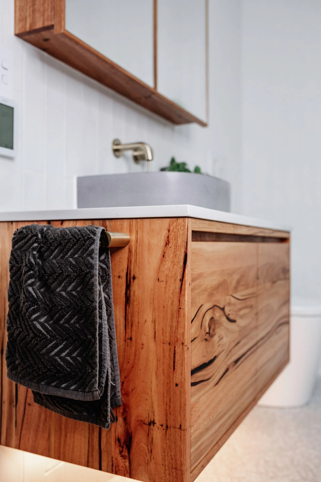 Close-up of a wooden cabinet with a black and gray patterned towel hanging from a gold towel bar in a bathroom. The bathroom has a white countertop and tile wall, with a round vessel sink and a gold faucet.