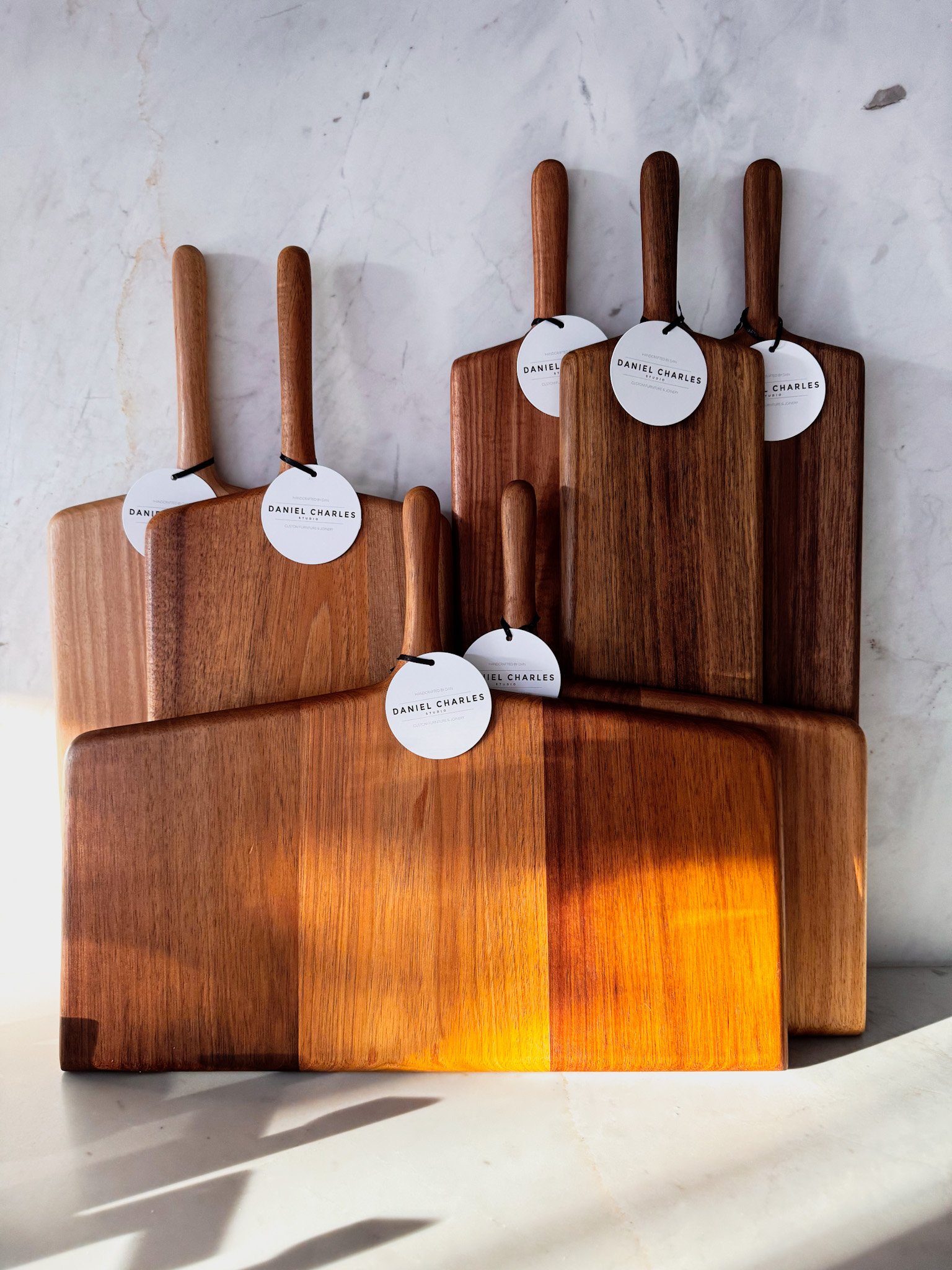 Three wooden cutting boards with handles, hanging tags labeled 'Daniel Charles Studio,' positioned on a marble or concrete wall.
