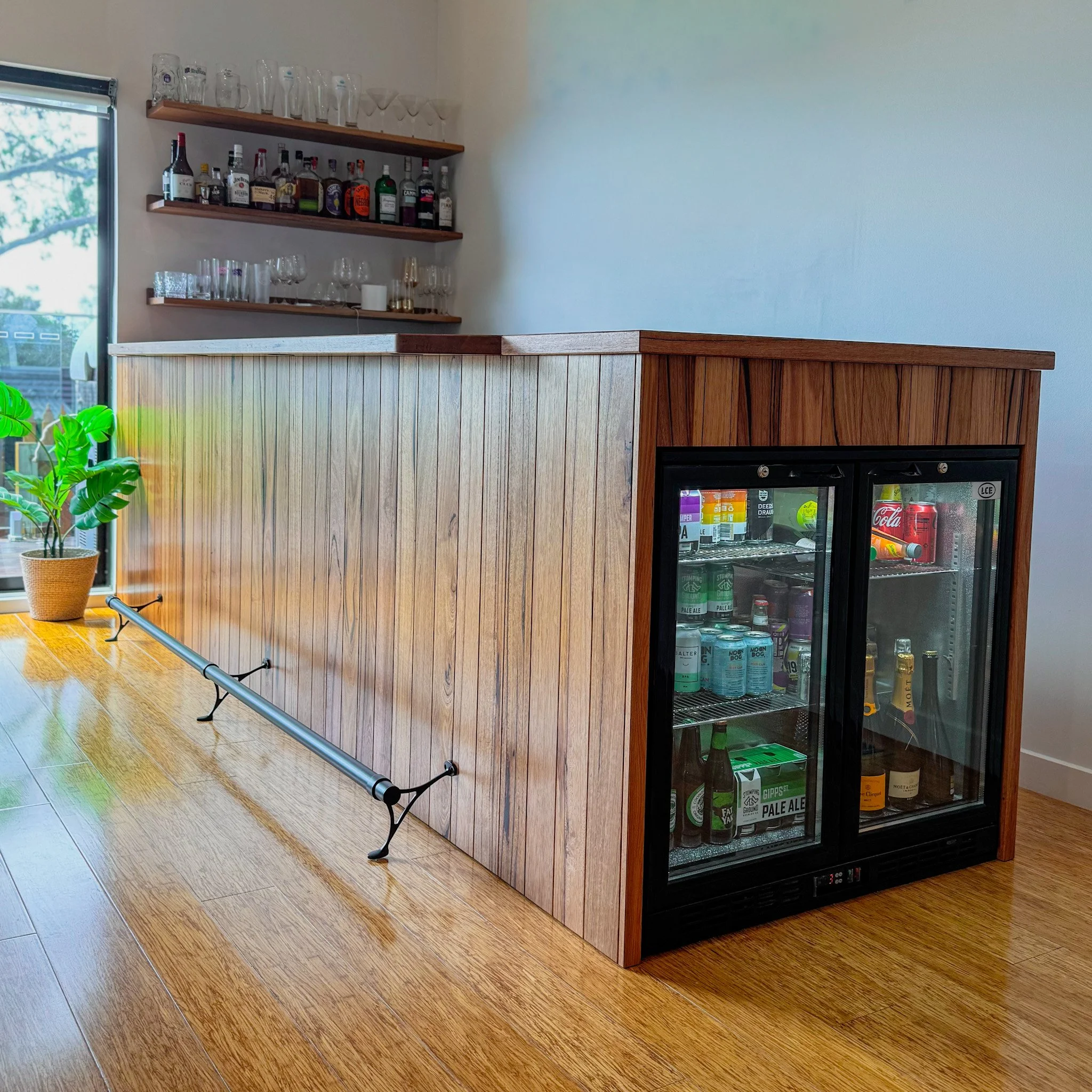 A bar area with two wooden shelves holding glassware and liquor bottles, a wooden counter, a potted plant, and a glass-front mini refrigerator filled with beverages, against a white wall and wooden floor.