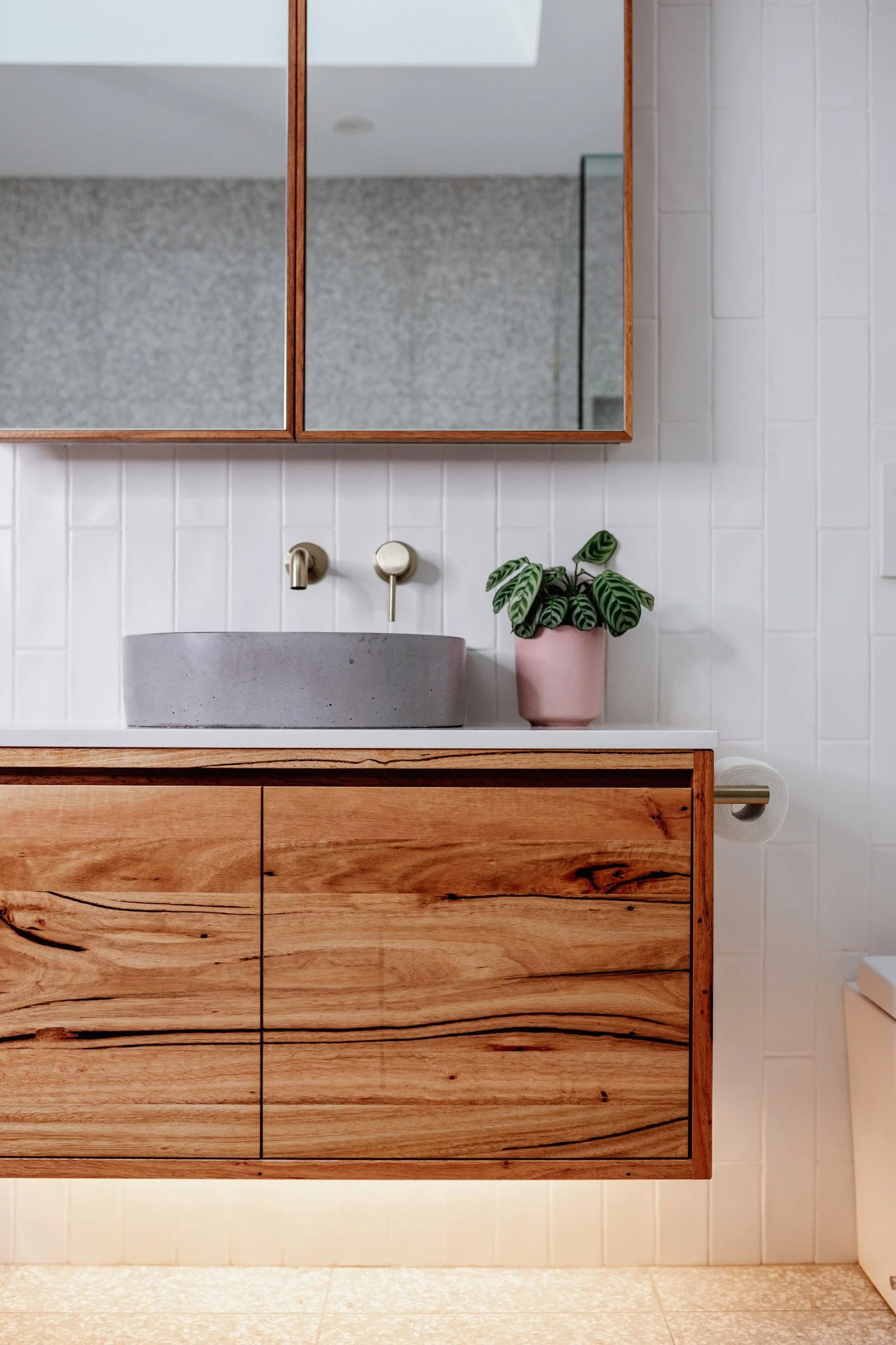A bathroom vanity with a wooden cabinet, a concrete sink, a wall-mounted faucet, a potted plant, a mirror with a wooden frame, white tiled walls, and a paper towel holder.