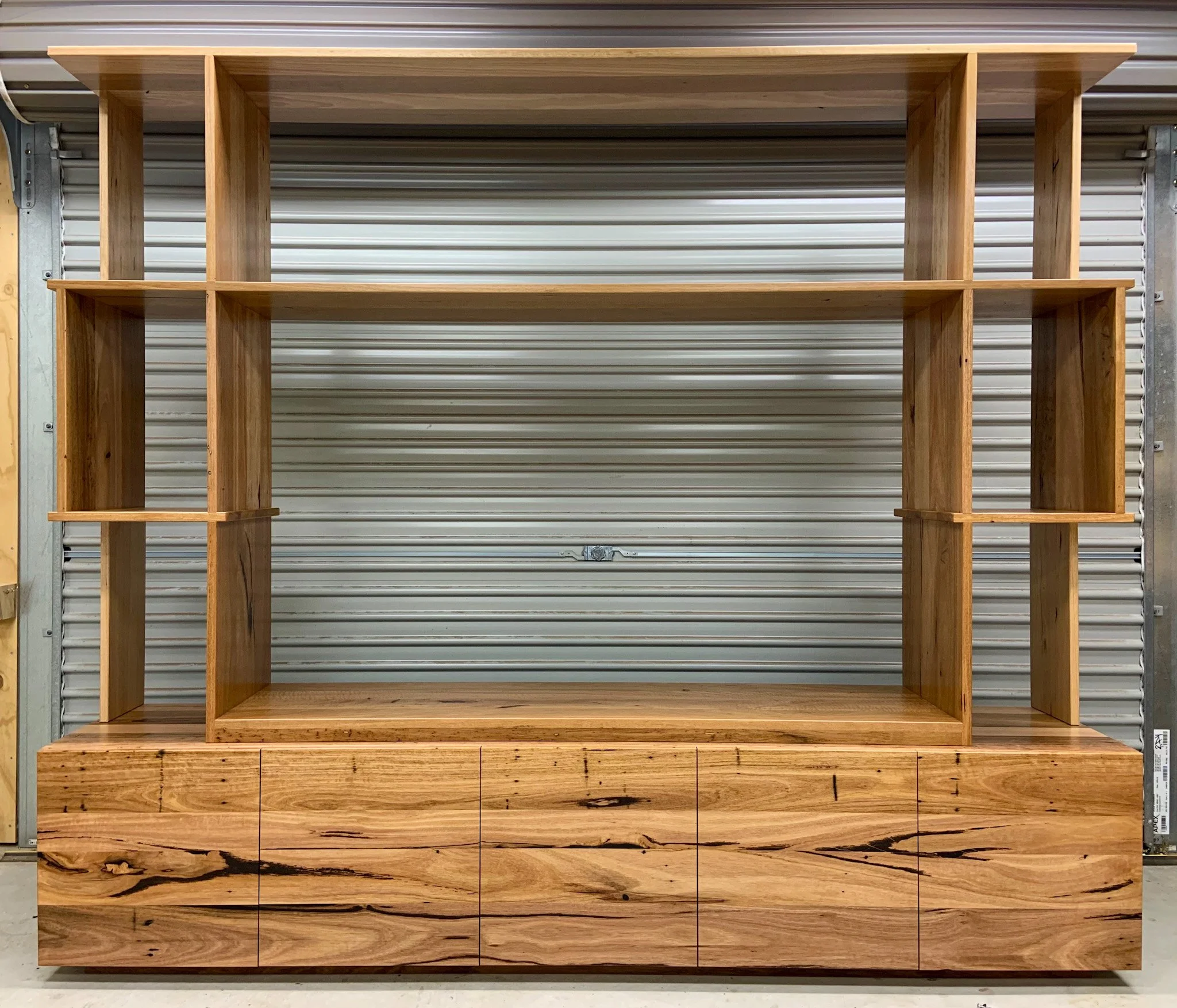 A large wooden bookshelf with multiple shelves, empty and made of light-colored, natural wood, stands against a metal roll-up door background.