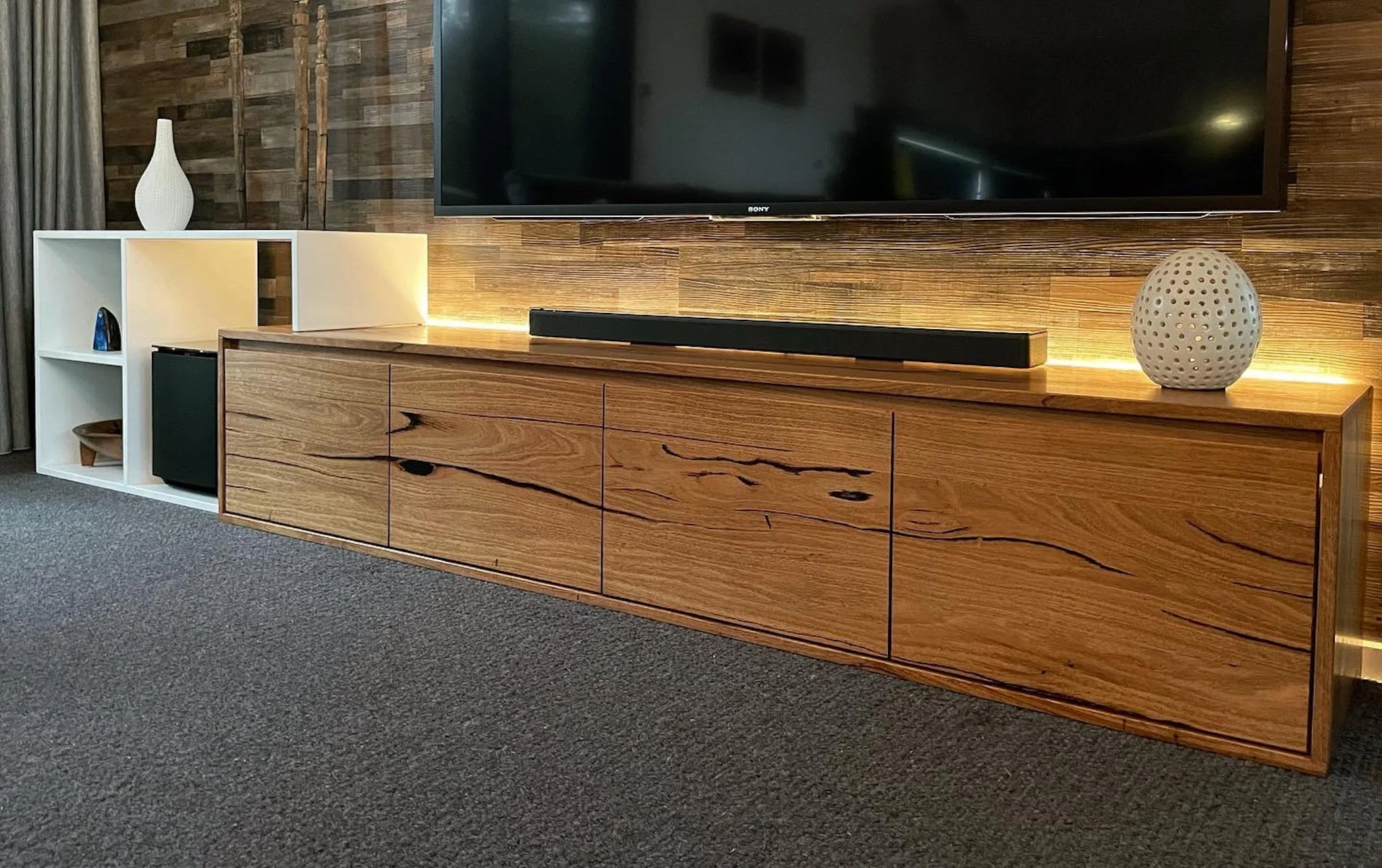 Living room entertainment area with a large flat-screen TV mounted on a wood-paneled wall. Below the TV is a wooden cabinet with a natural finish and black accents. To the left, a white storage unit with open shelves and a black speaker. Decorative i