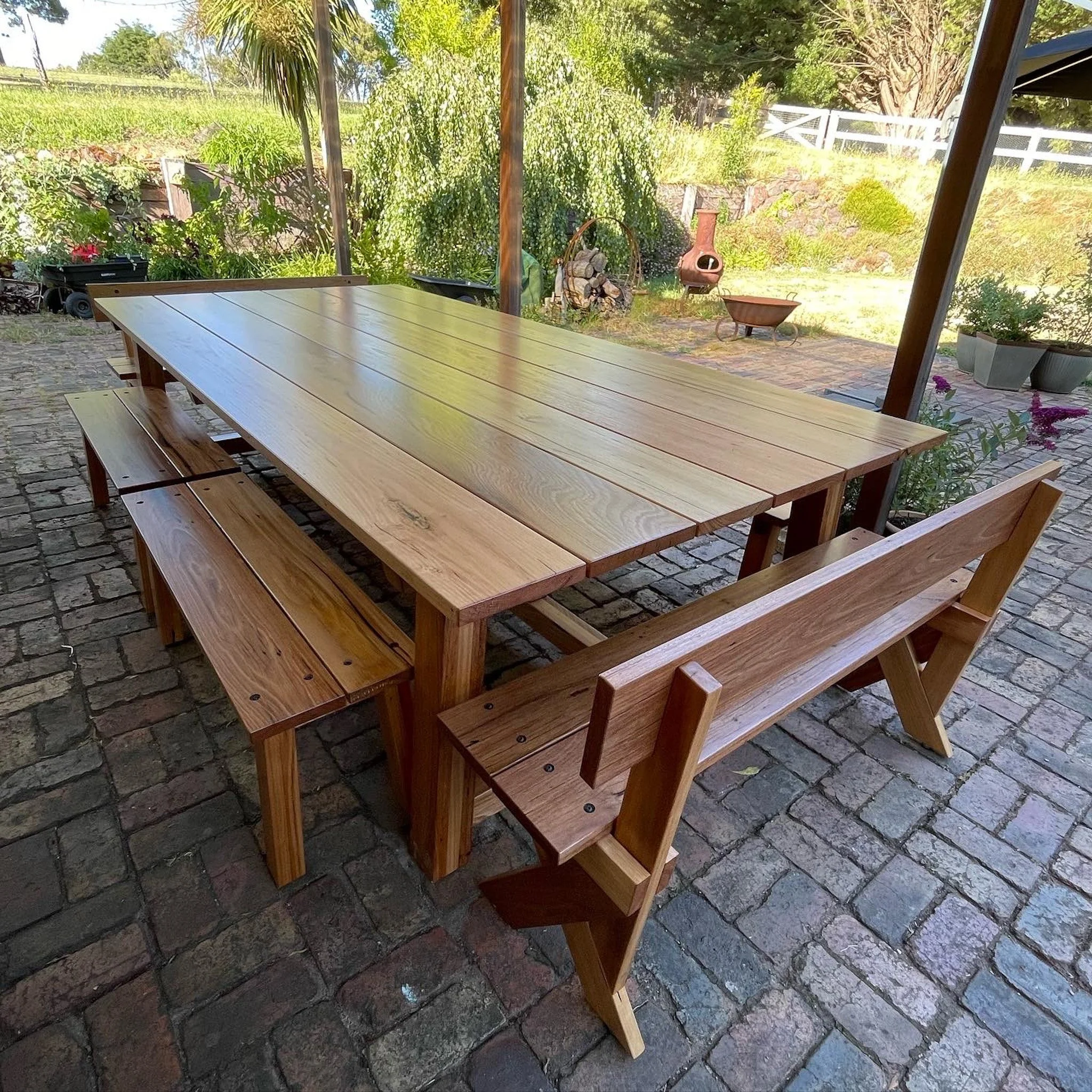 A large wooden outdoor dining table with matching benches on a stone patio, with a lush garden and trees in the background.
